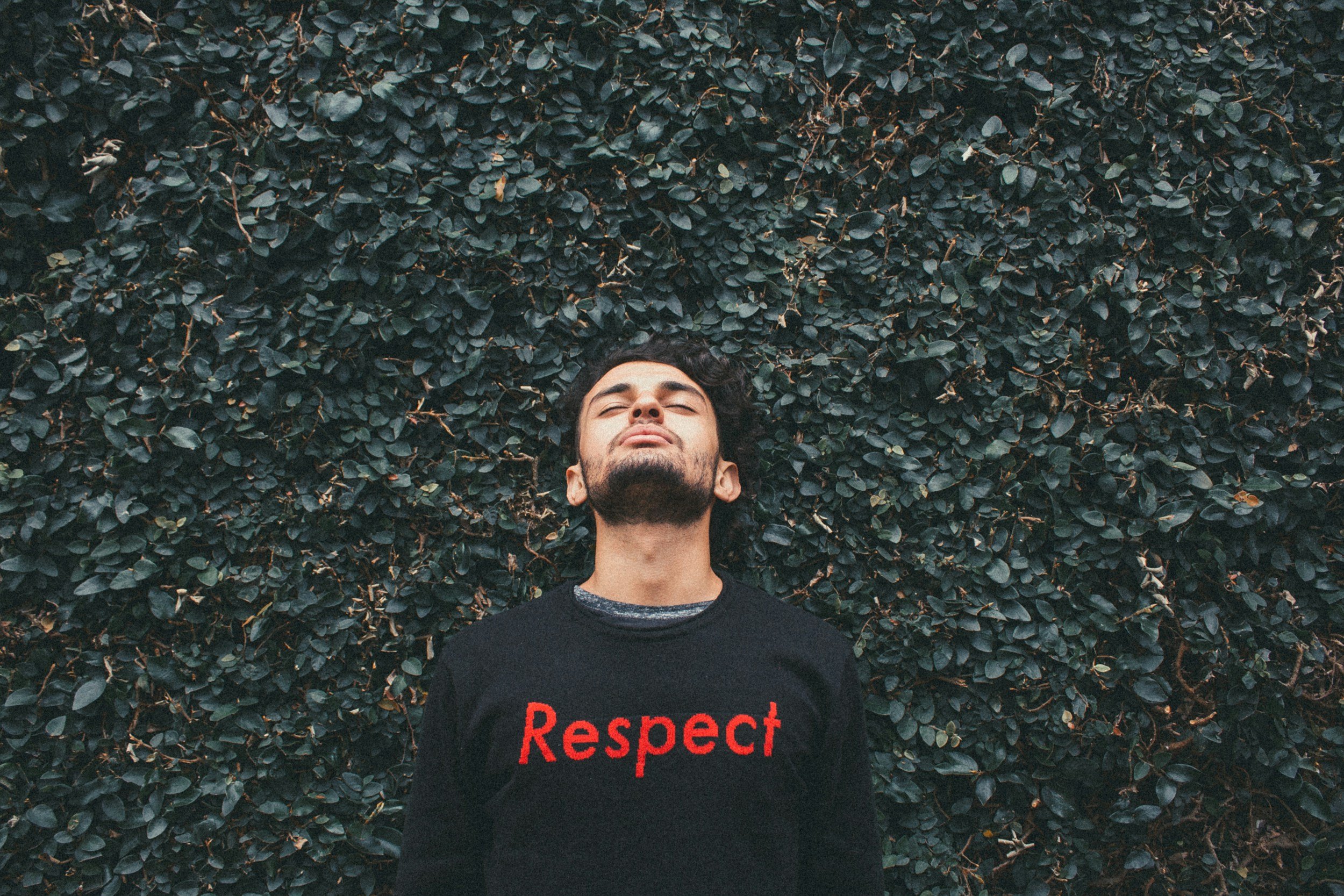 Young man standing in front of a dense green bush with closed eyes and a peaceful expression, wearing a black sweater with the word 'Respect' in red letters.
