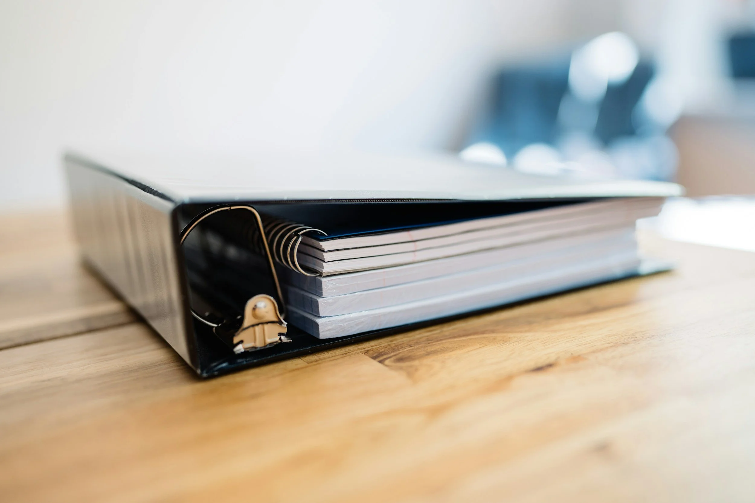 A black binder with a gold paperclip on top, holding several notebooks, on a wooden table.
