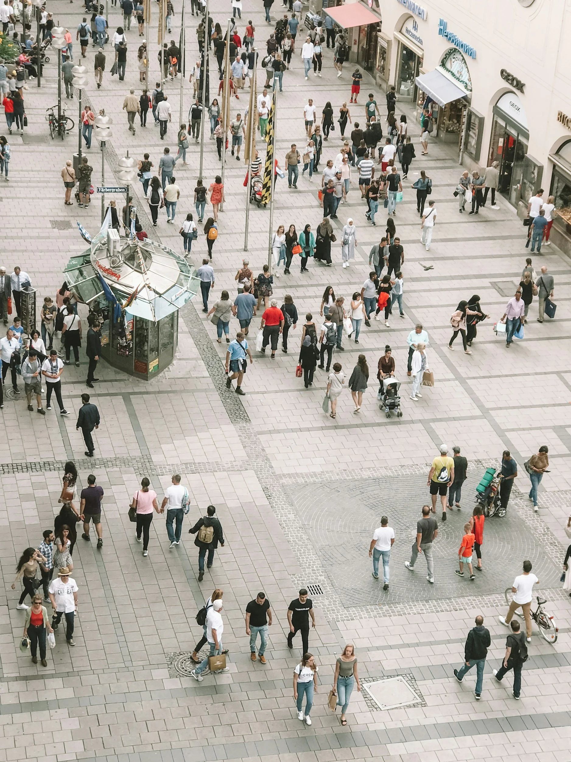 A busy city plaza filled with pedestrians walking in various directions. The area has shops, street vendors, and flag poles, with some people pushing strollers and cycling.