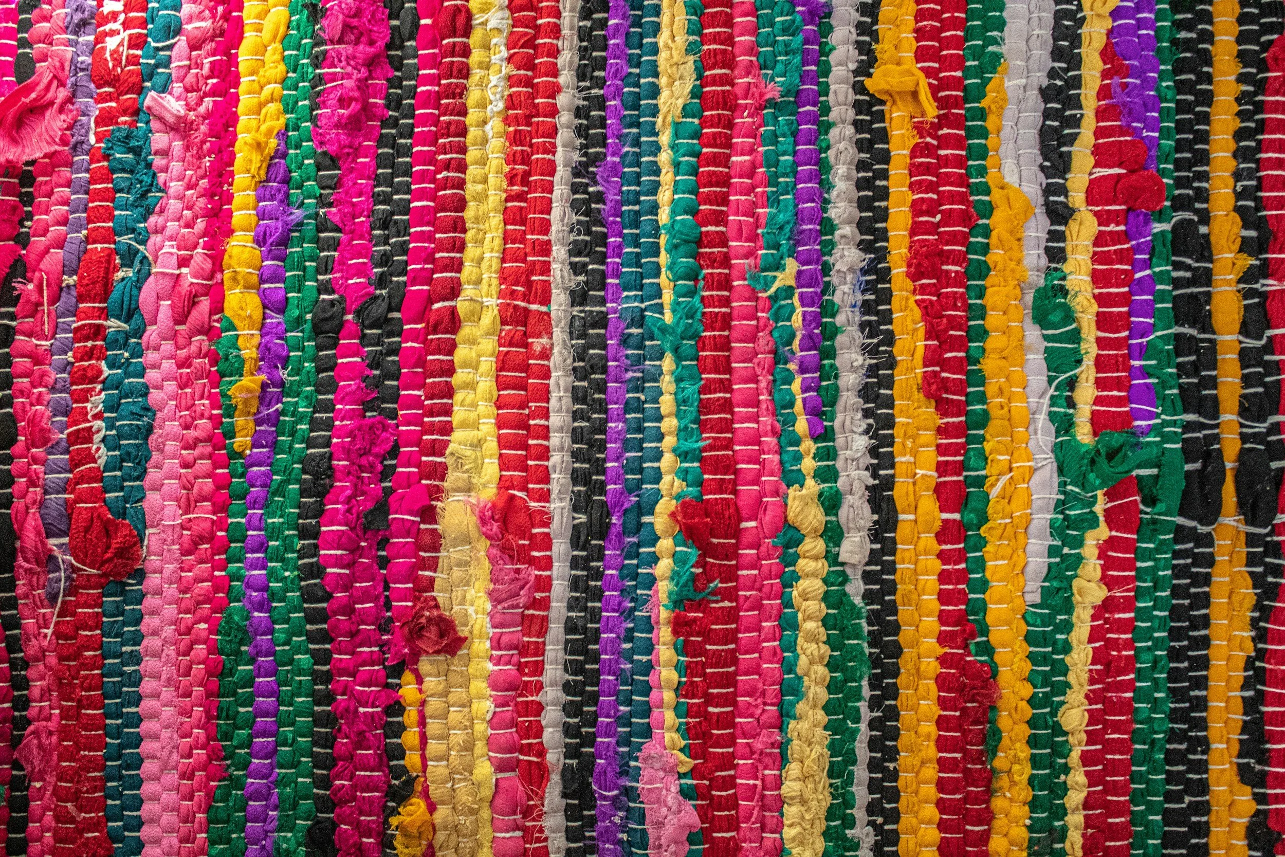 Colorful woven fabric with various strips of yarn in pink, purple, yellow, green, black, red, orange, and white.