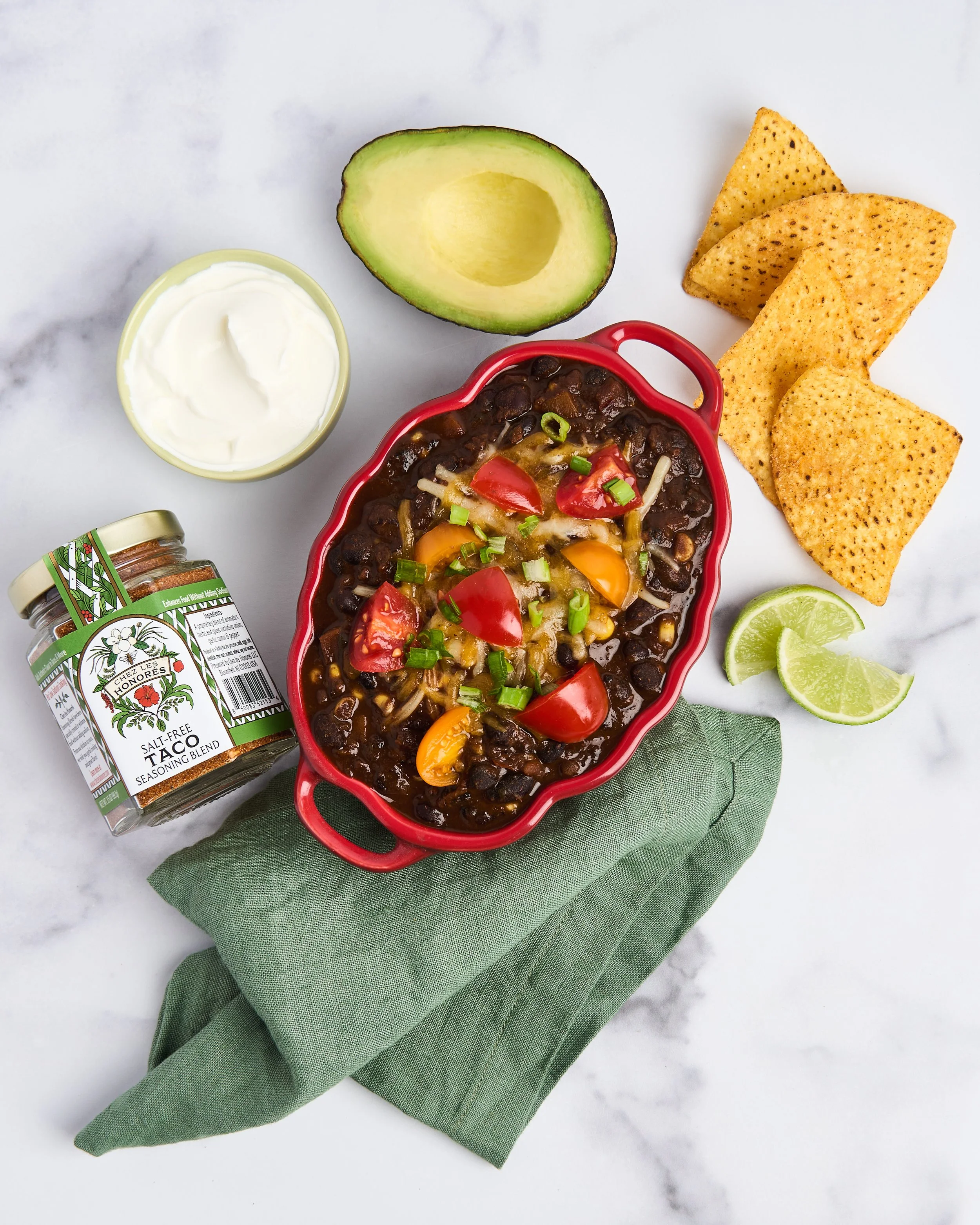 A bowl of chili with tomatoes, chili, and cheese topped with green onions, served with lime wedges, tortilla chips, avocado half, sour cream, and taco seasoning on a white marble surface.