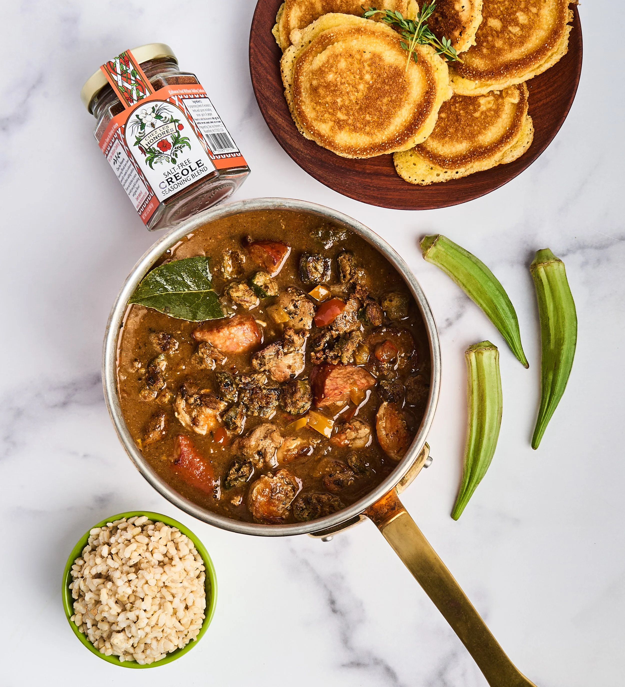 A bowl of beef stew with vegetables, a small green bowl of rice, a wooden plate of fried pancake-like food, a jar of seasoning, and green okra on a white marble surface.