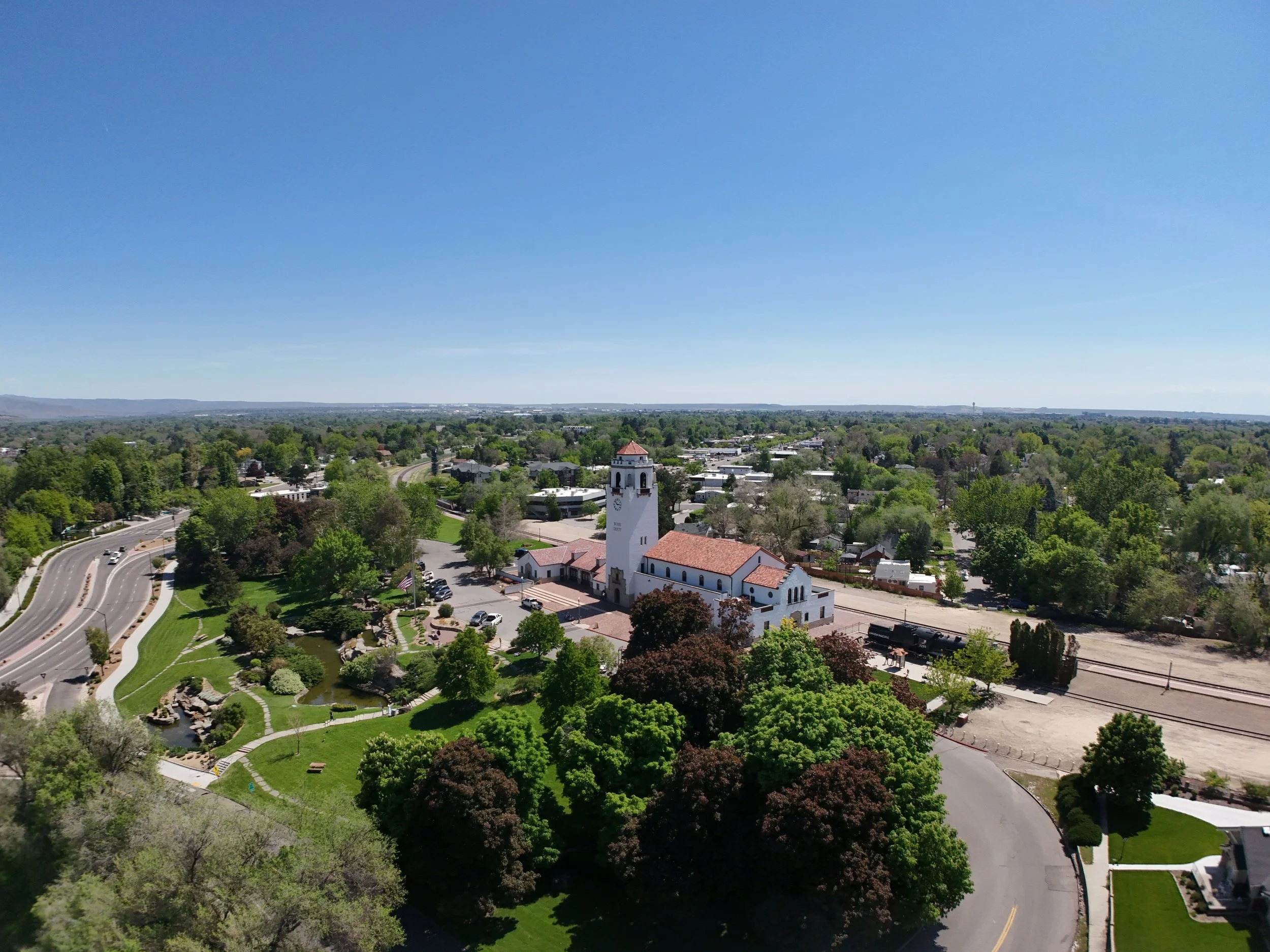 Aerial view of a town with a white church, surrounded by green trees, roads, houses, and a park with a pond, under a clear blue sky.