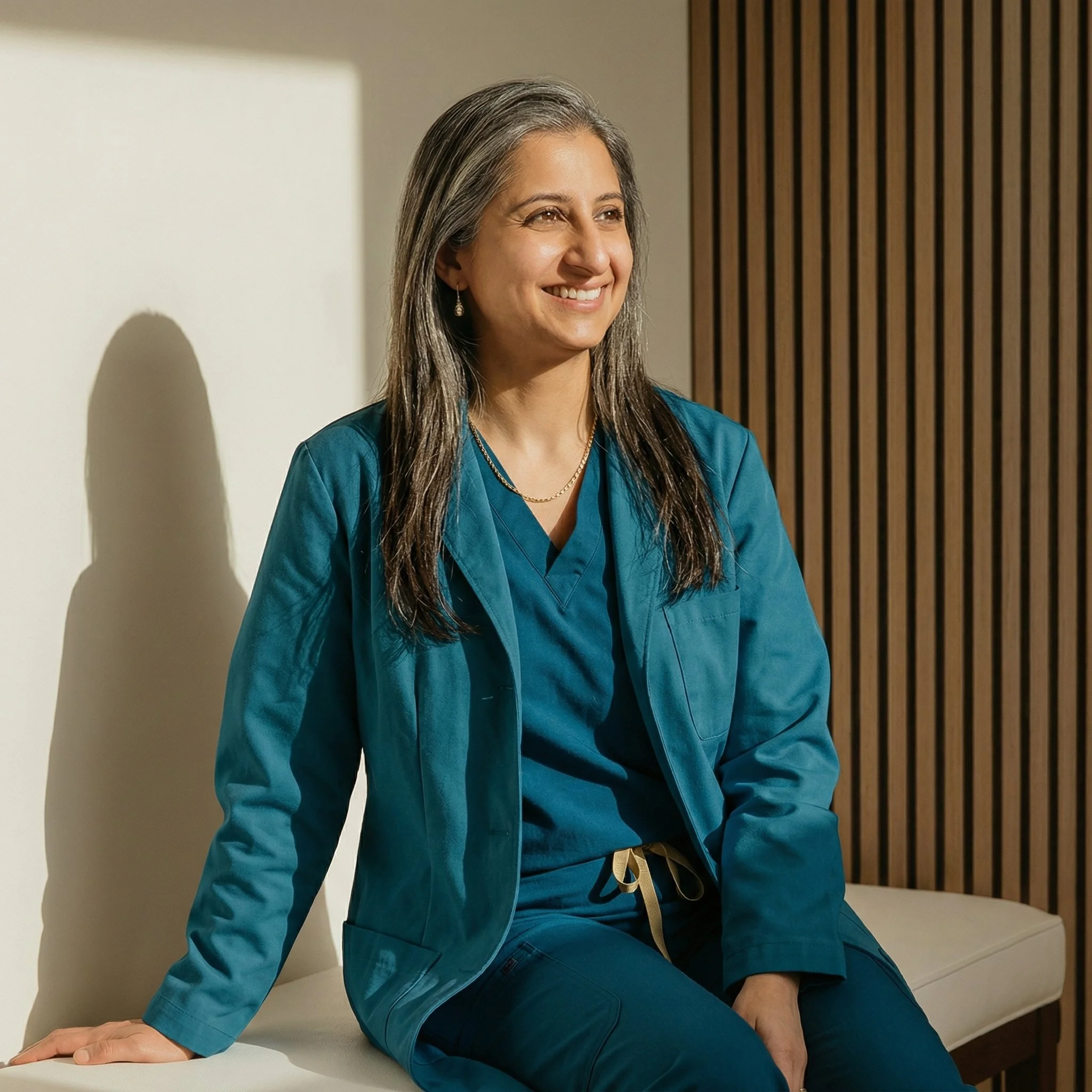 A woman with long gray hair wearing blue medical scrubs, sitting and smiling indoors with wooden wall paneling in the background.