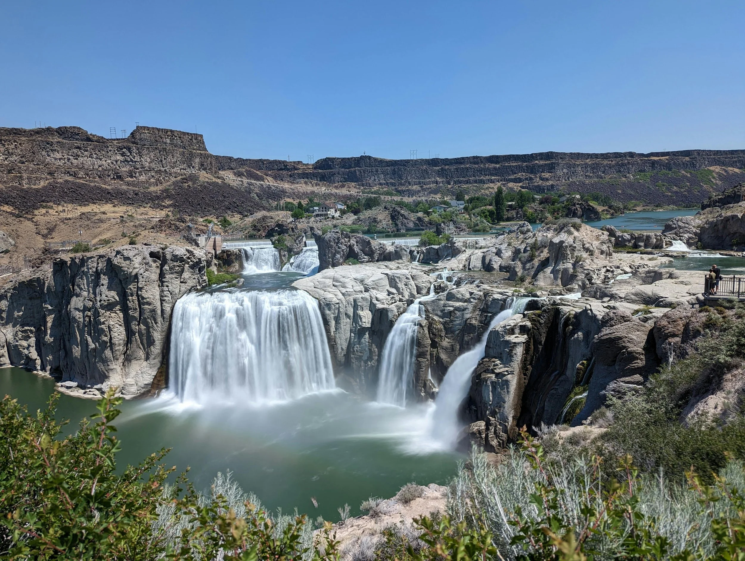 A scenic view of a waterfall cascading over rocks with a river flowing in a canyon, surrounded by arid, rocky hills and sparse vegetation under a clear blue sky.