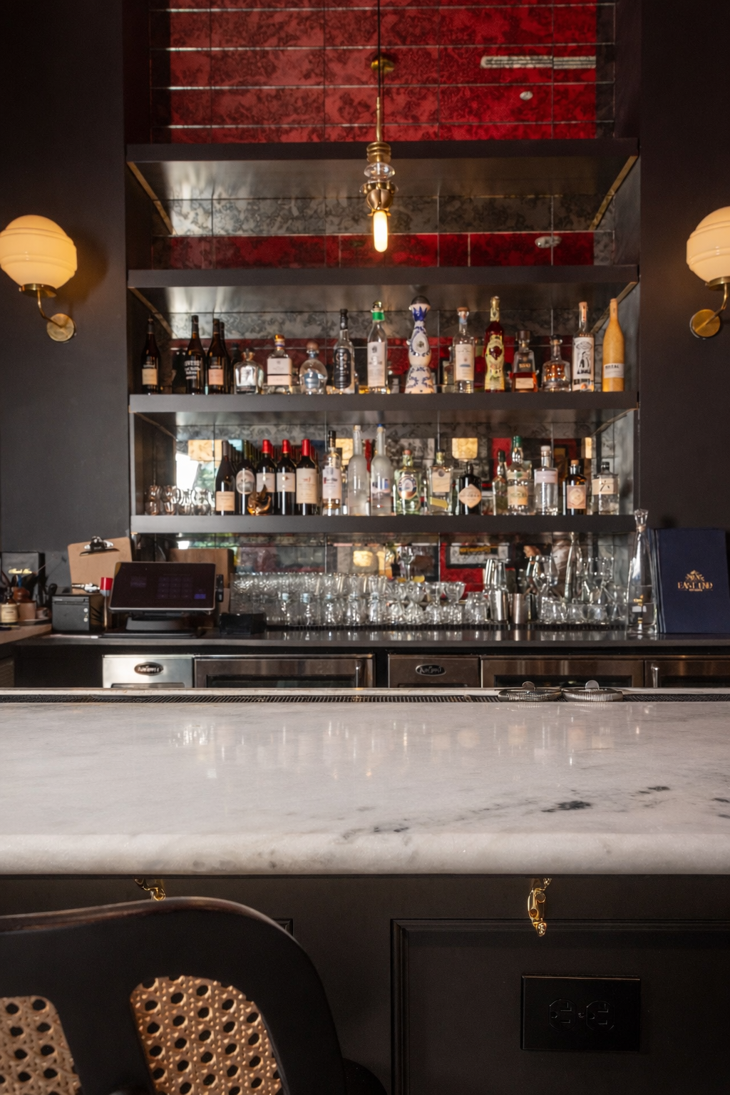Bar with shelves of liquor bottles, glasses, and bar equipment behind a marble countertop, with red walls and black accents.