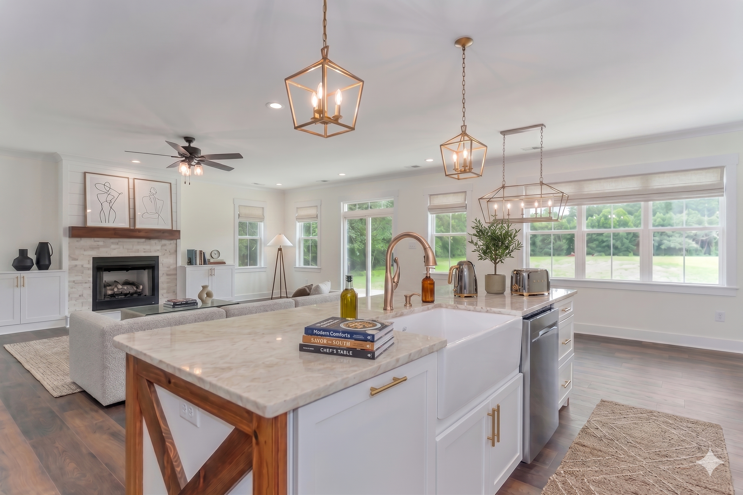 Bright, modern kitchen with white cabinets and marble countertops, featuring pendant lighting, large windows with views of greenery, and a living area with a fireplace and abstract art on the wall.