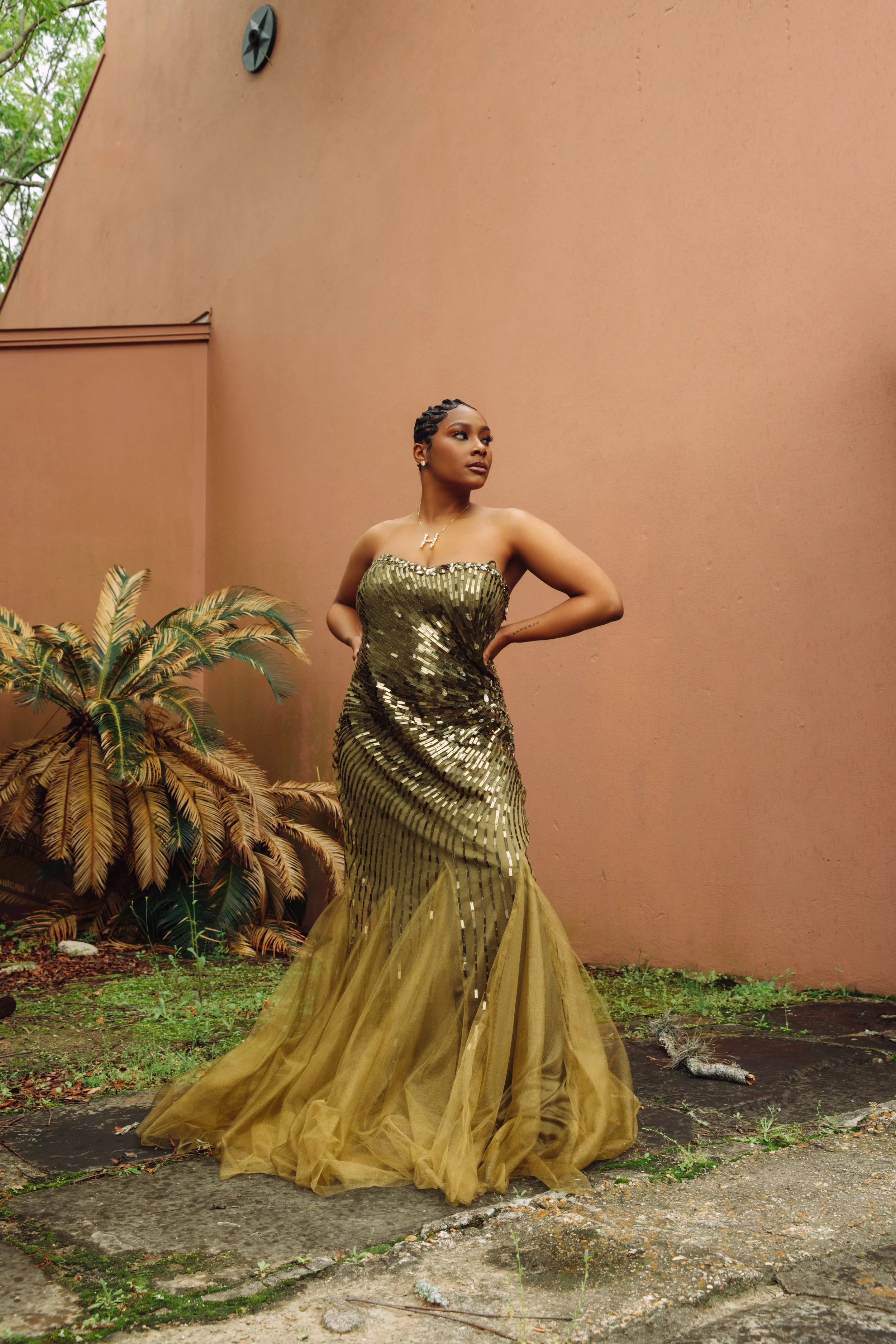 A woman in a strapless, gold, sequin gown with a tulle train standing outdoors against a pink wall with plants behind her.