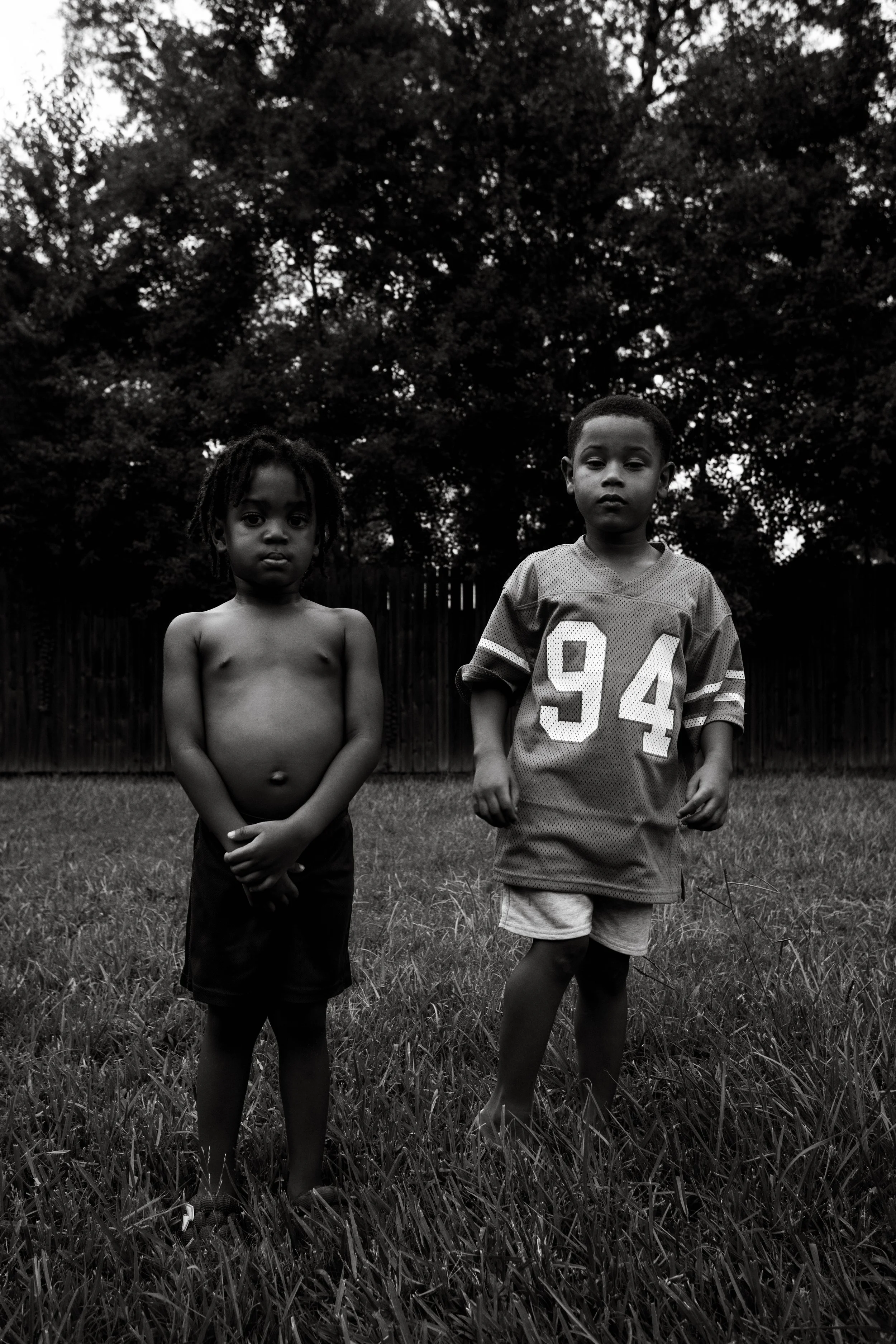 Two young boys standing in a grassy yard with a fence and trees in the background, one shirtless and the other wearing a sports jersey, in black and white.