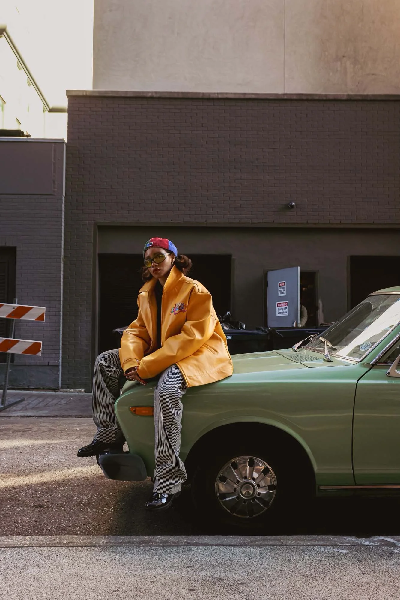 A woman wearing a yellow jacket, plaid pants, and a red, blue, and white baseball cap, sitting on the hood of a green vintage car parked on an urban street.