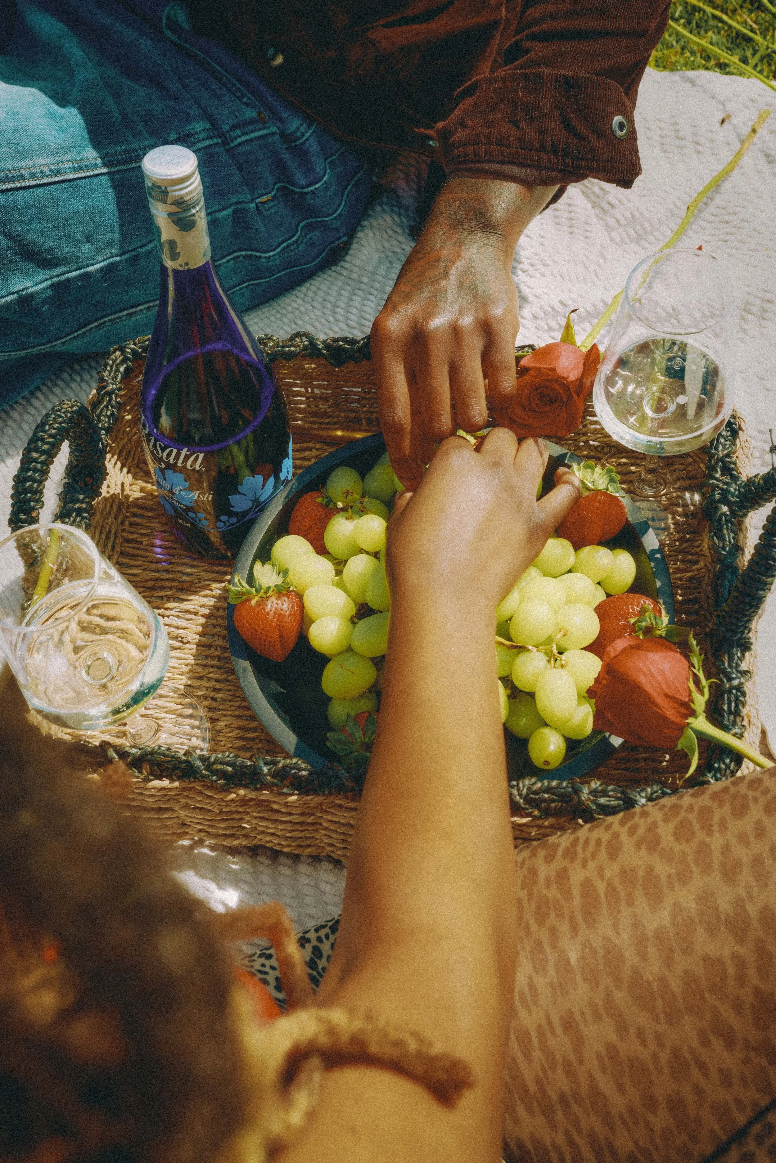 People sharing a picnic with grapes, strawberries, roses, wine, and glass cups on a wicker tray.