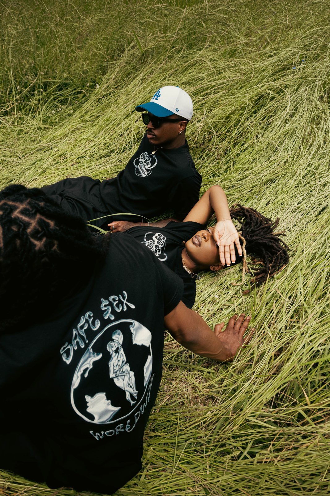 Two people are lying in tall grass, one with dreadlocks and the other wearing a cap and sunglasses, both wearing black T-shirts with a graphic logo.