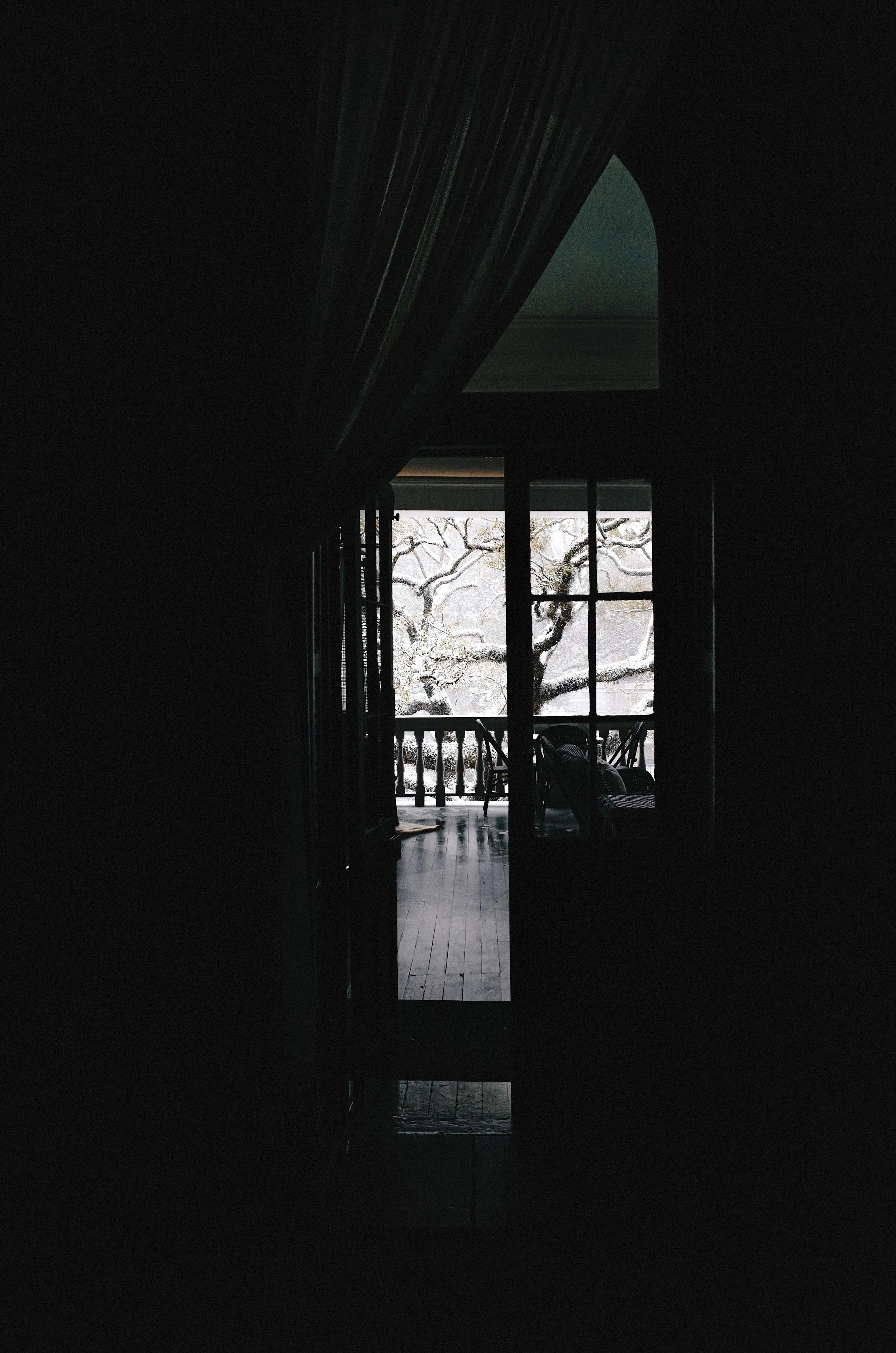 Inside view of a dark room looking out through an open door to a balcony with outdoor seating, trees, and overcast sky in the background.