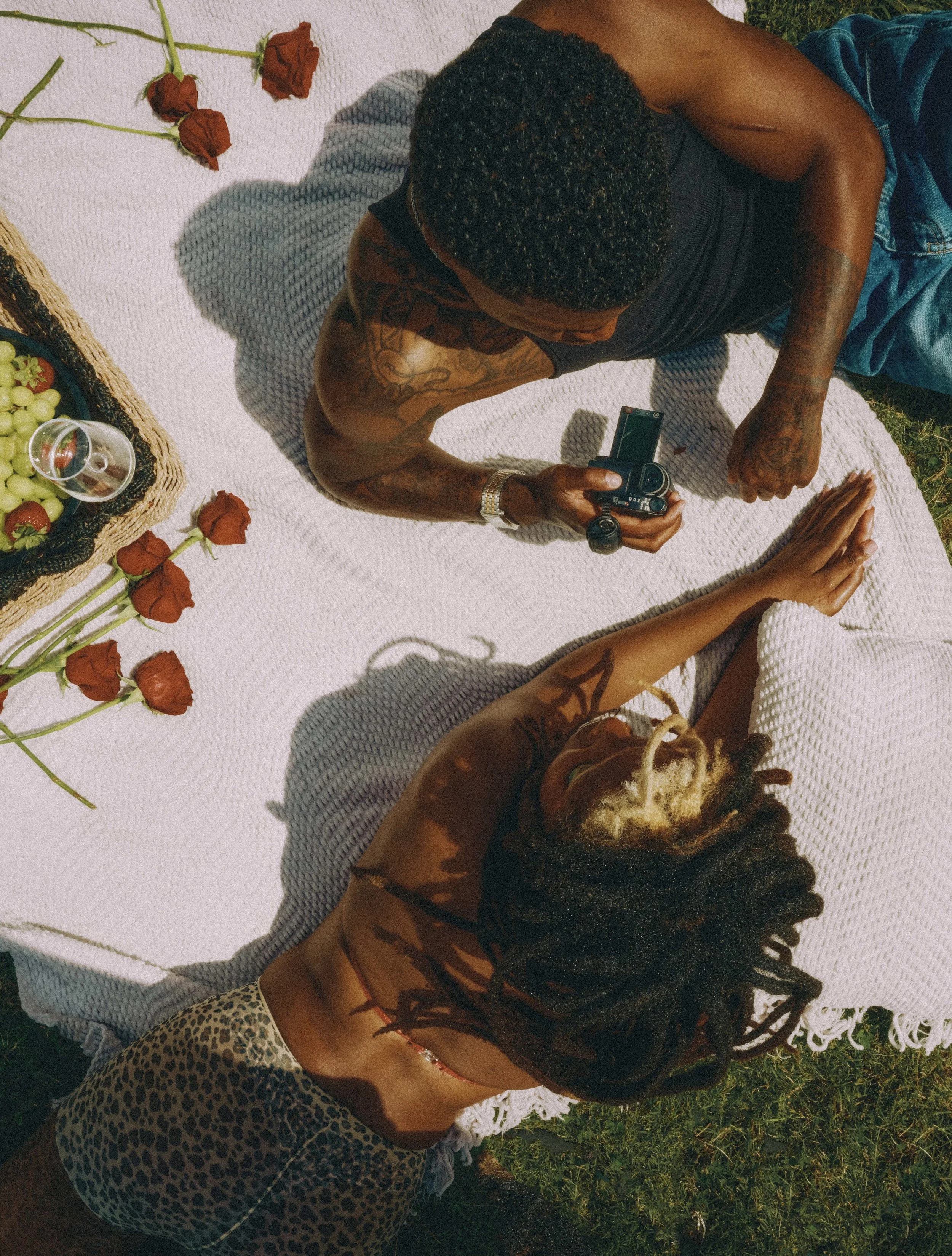 A man and woman lie on a blanket outdoors, with the man holding a camera. There are red roses, a basket of grapes, and a glass of wine on the blanket.
