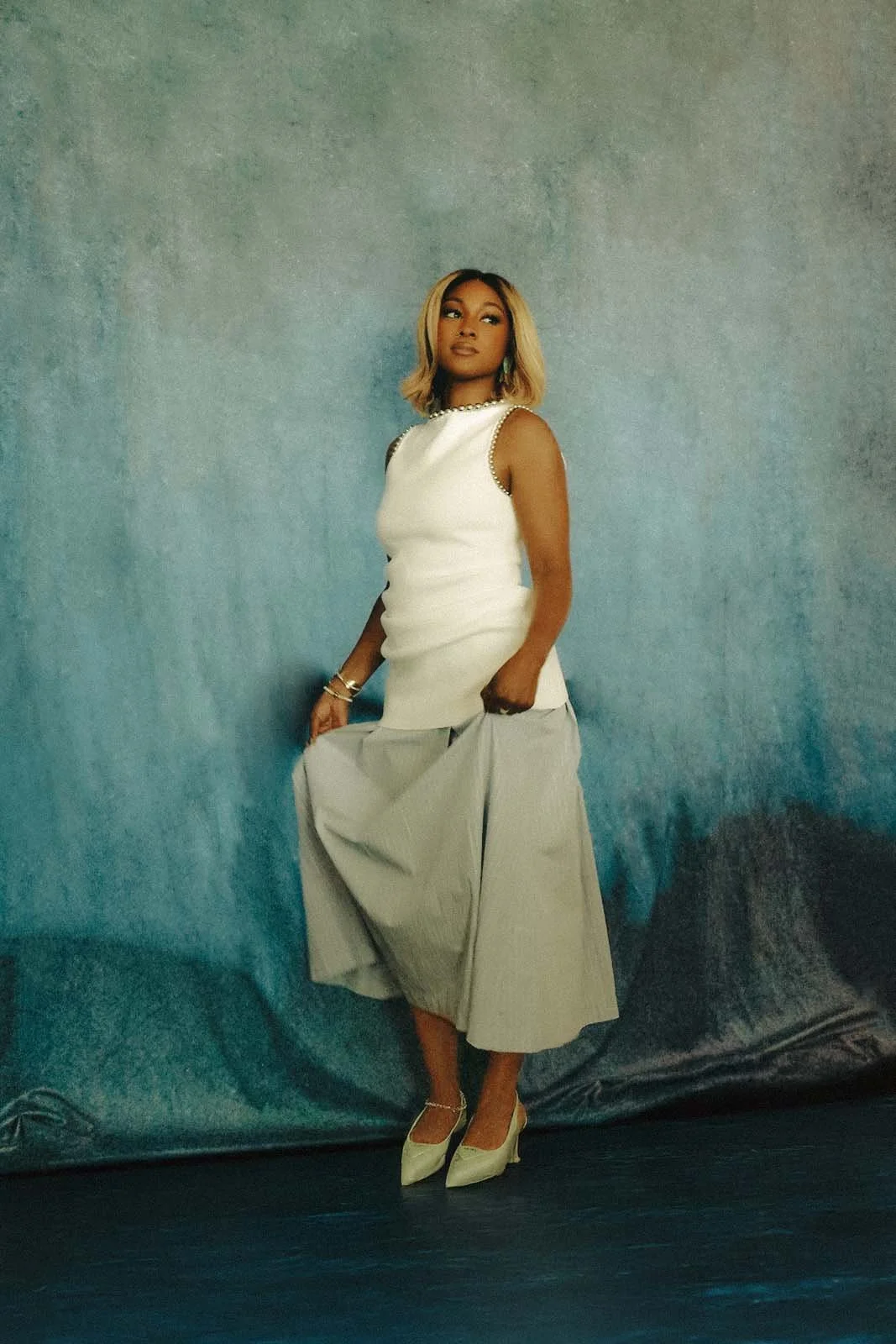 A woman in a sleeveless white top and a gray wide-leg skirt, holding the skirt up slightly, standing against a textured blue background. She has shoulder-length blond hair and is wearing gold jewelry and white high heels.