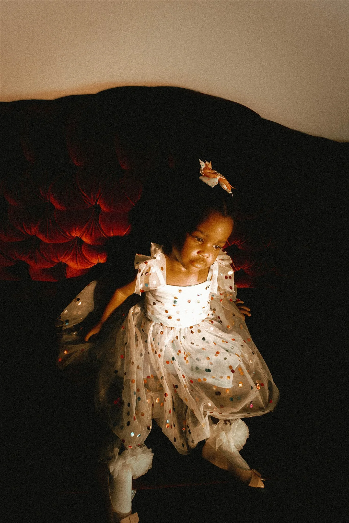 A young girl in a white dress with multicolored polka dots sits on a red velvet tufted couch, looking downward.