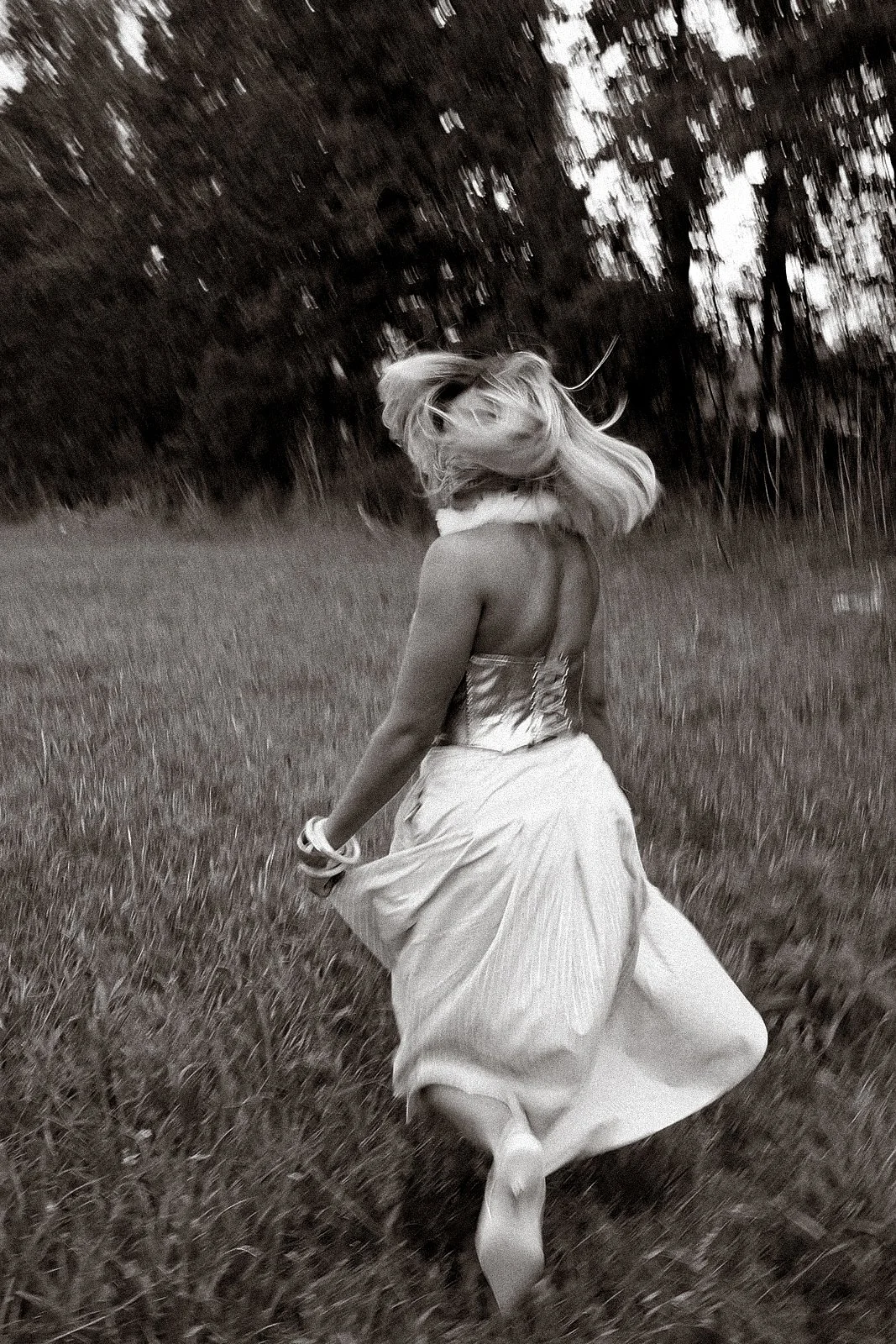 A woman with long blonde hair running through a grassy field, wearing a strapless dress and jewelry, with trees in the background.