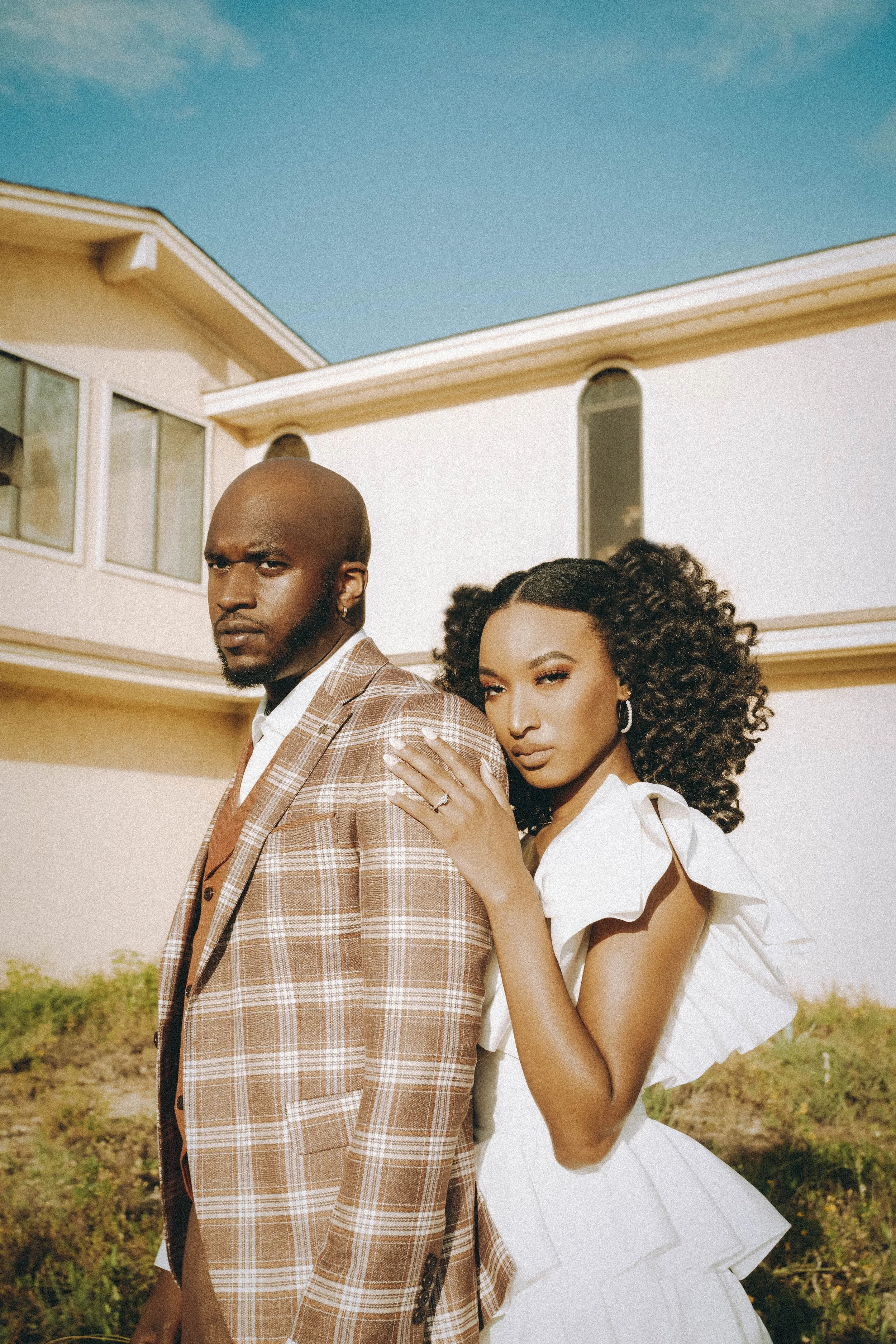 A man and woman standing outdoors in front of a white house under a clear blue sky. The man is wearing a brown plaid suit and the woman is wearing a white dress. The woman has her hand on the man's shoulder, and they both look serious.