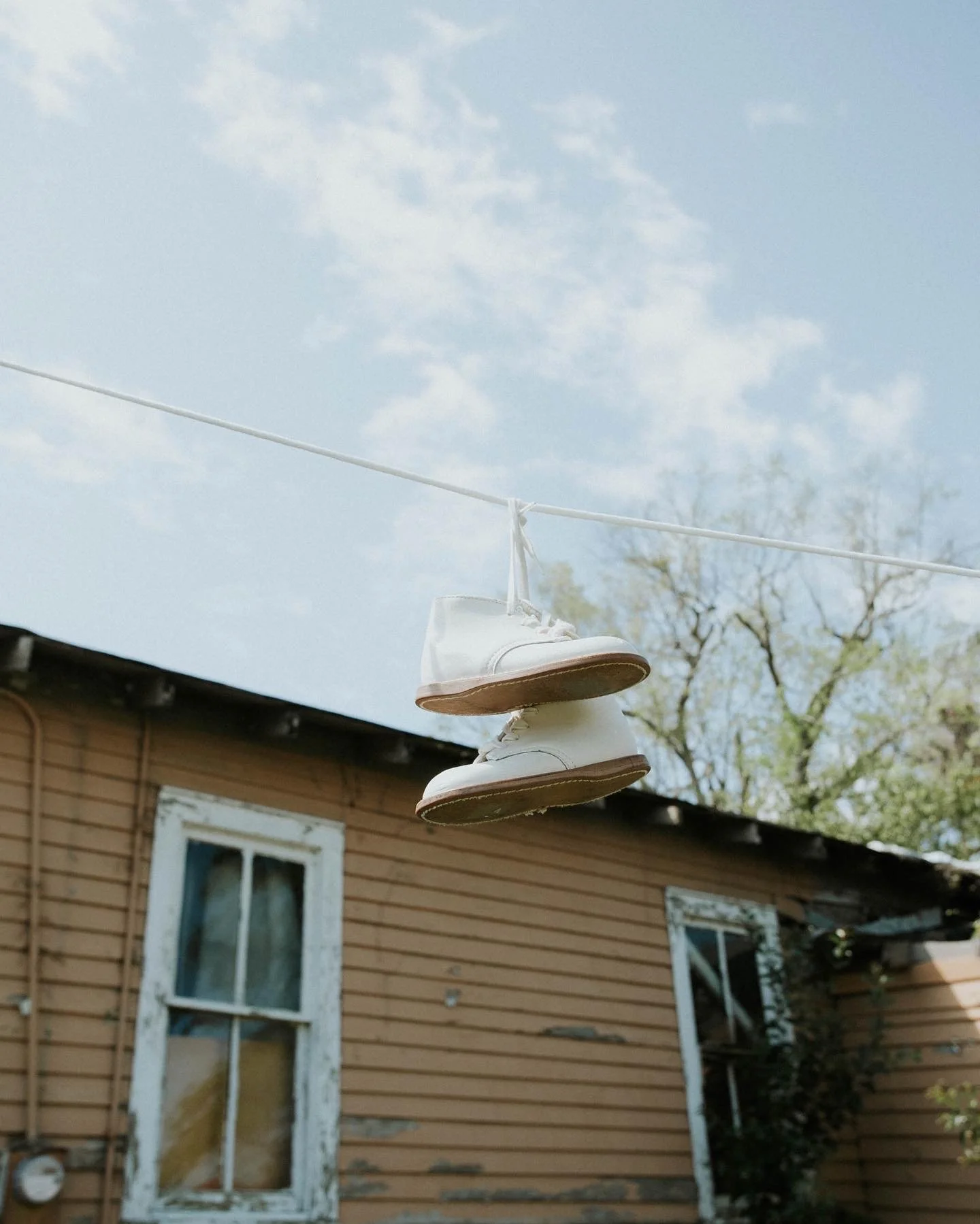 A pair of white sneakers hanging from a clothesline outside a house with weathered siding, a window, and trees in the background.