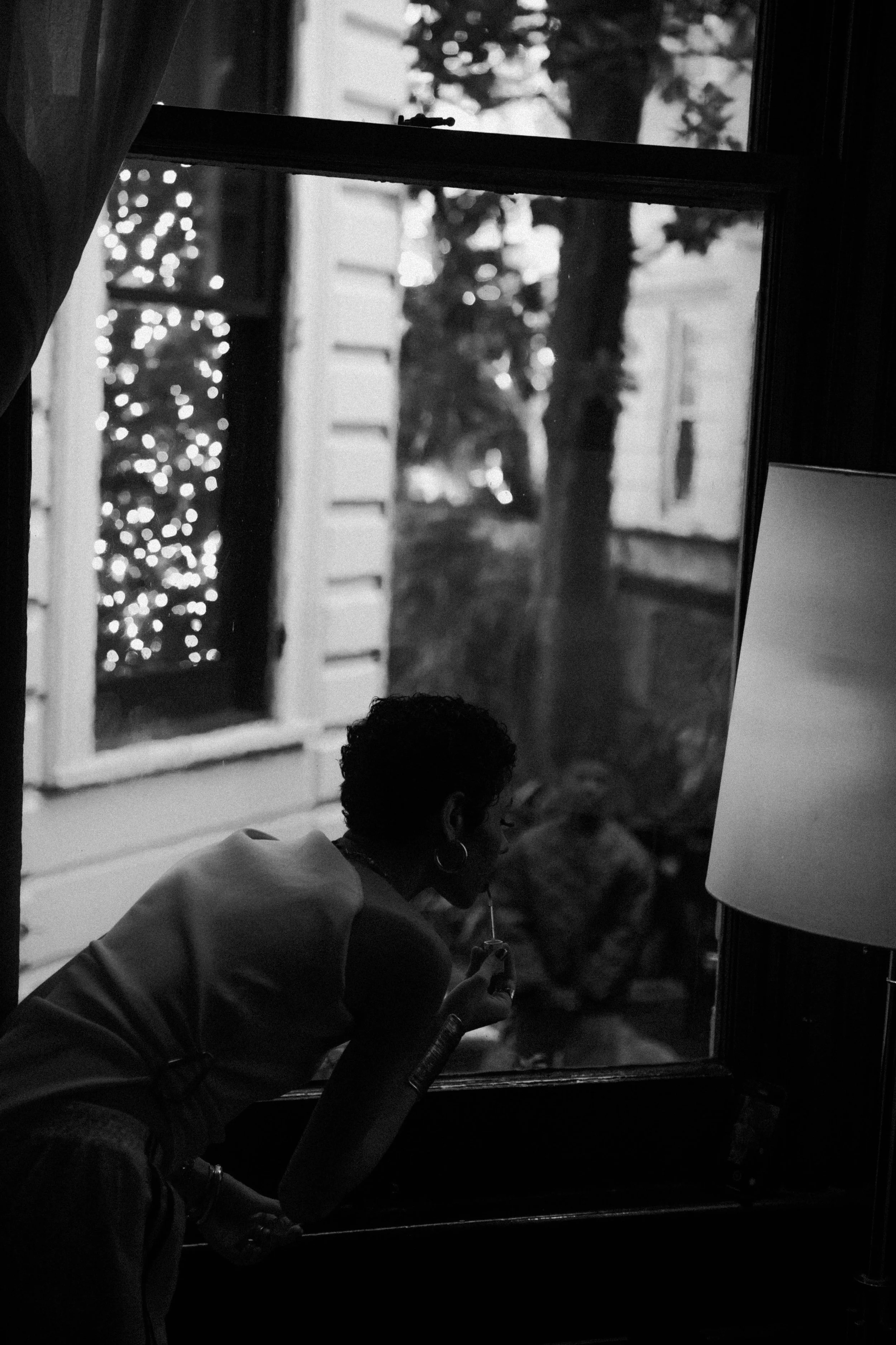 A woman leaning through a window and smoking an e-cigarette, with a Christmas tree with lights outside in the background.