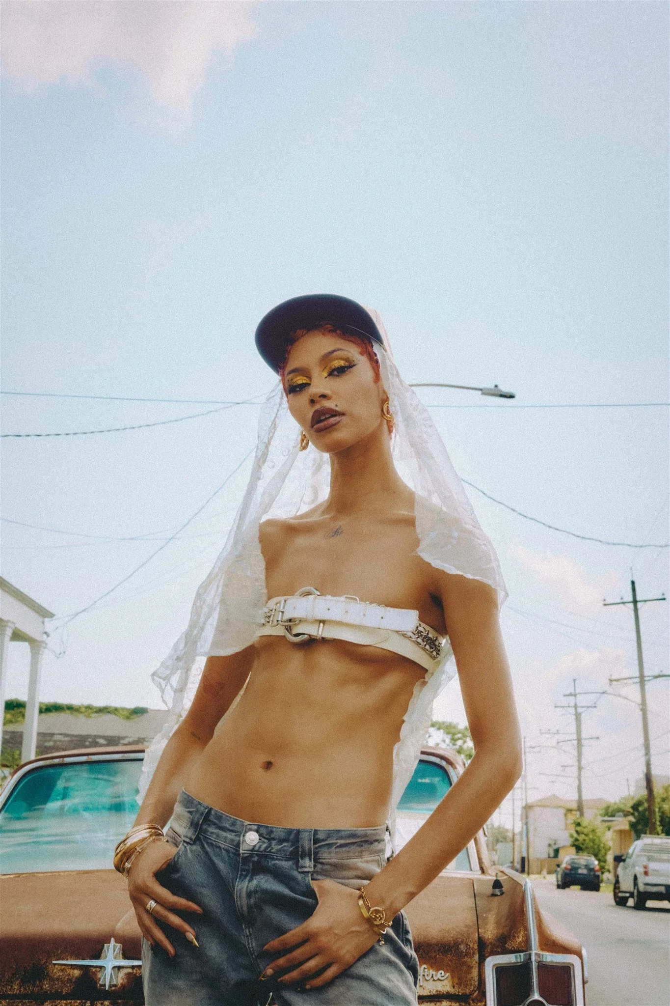 A person with short red hair and yellow makeup standing in front of a rusty vintage car on a street, wearing a black cap, a white strapless top with a belt, and gray jeans, with a veil draped over their head.