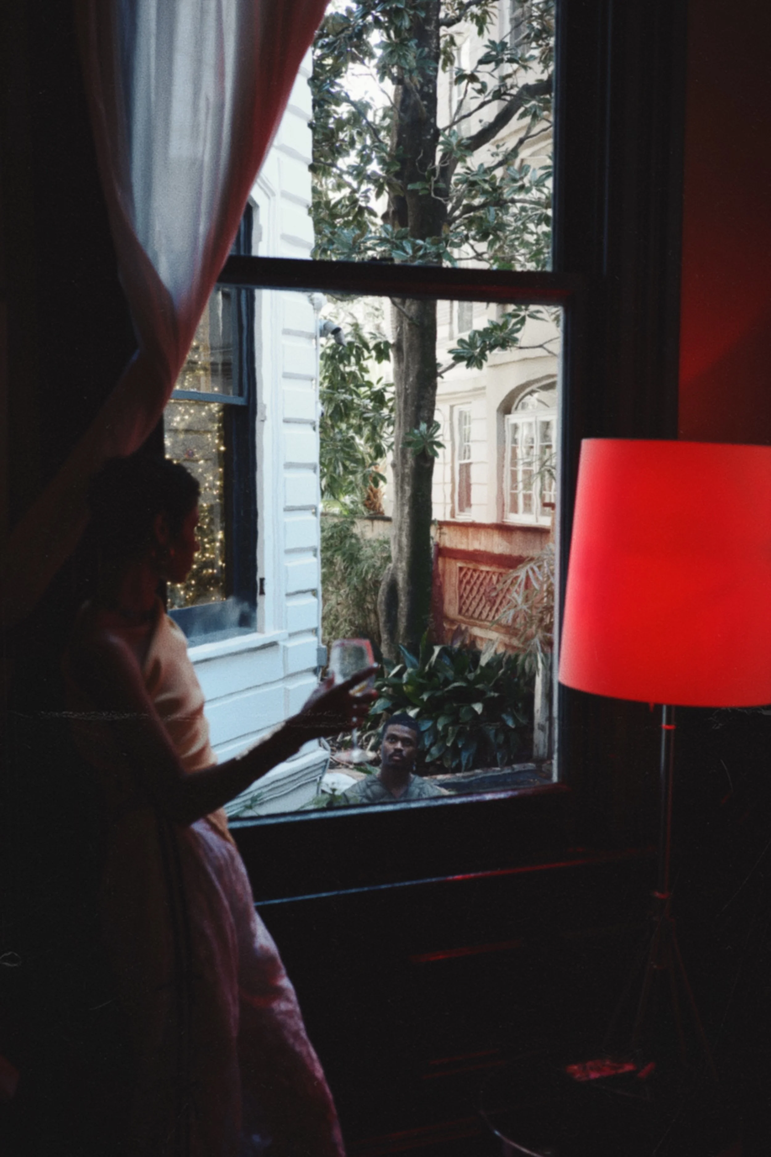 A woman indoors standing near a window, holding a wine glass, with a man outside visible through the window looking in. There's a large red lampshade on a lamp beside her.