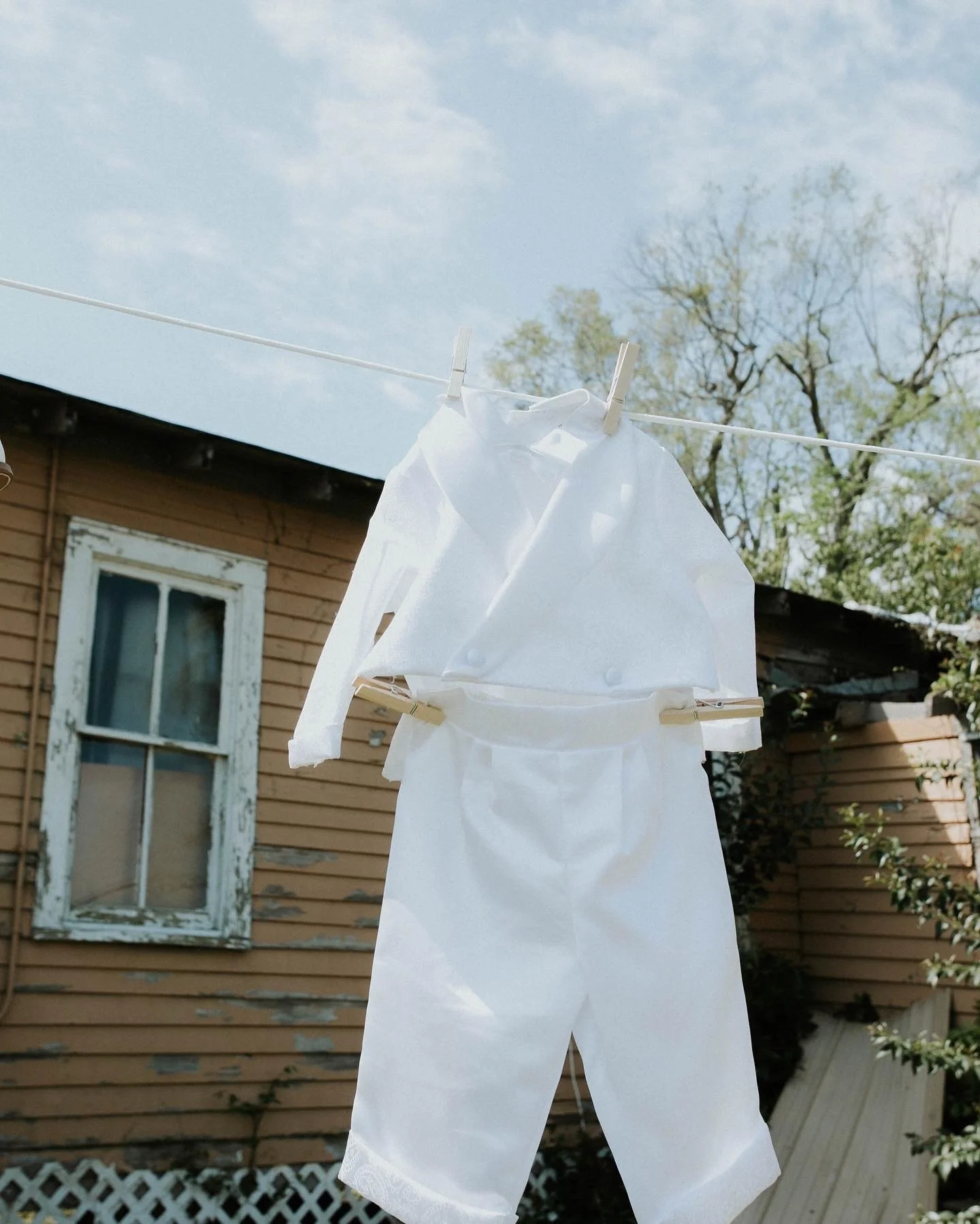 White baby clothes hanging on a clothesline outside in front of a house with a wooden exterior and a window with peeling white paint.