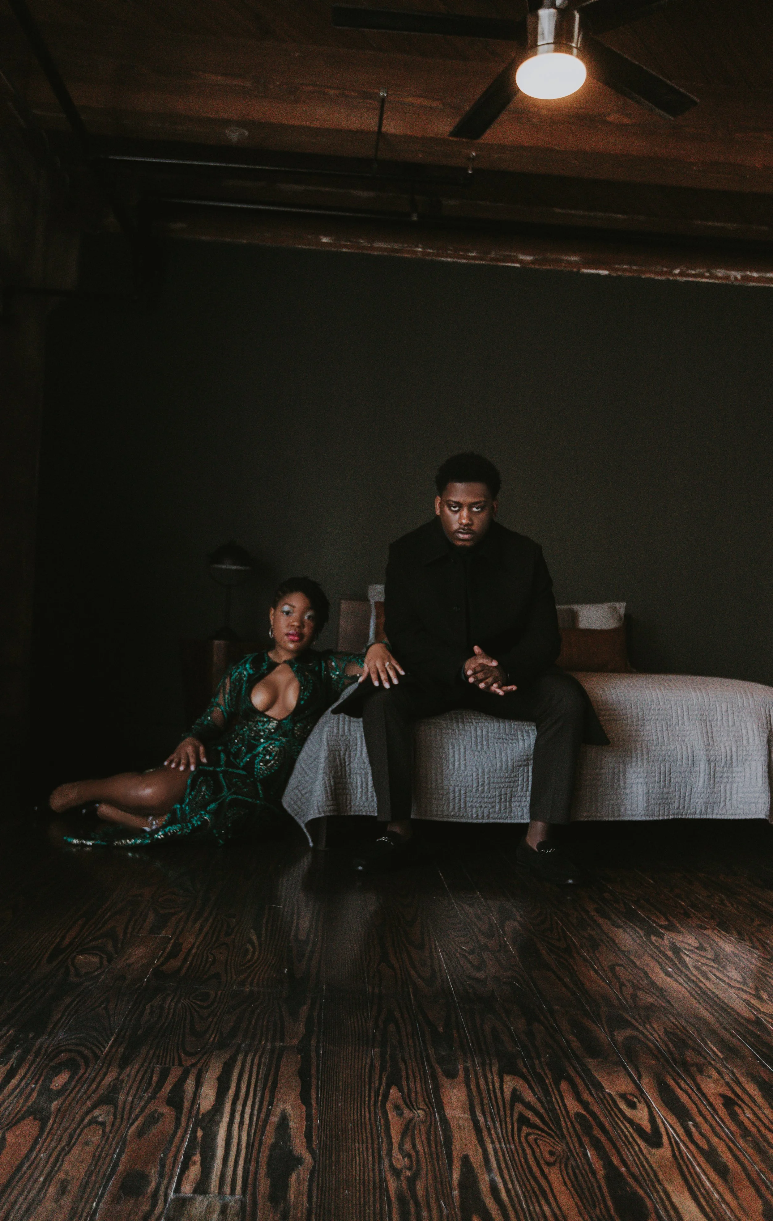 A woman in a green and black dress lying on the dark wooden floor next to a man in black sitting on a bed in a dimly lit room with dark walls and wooden ceiling.