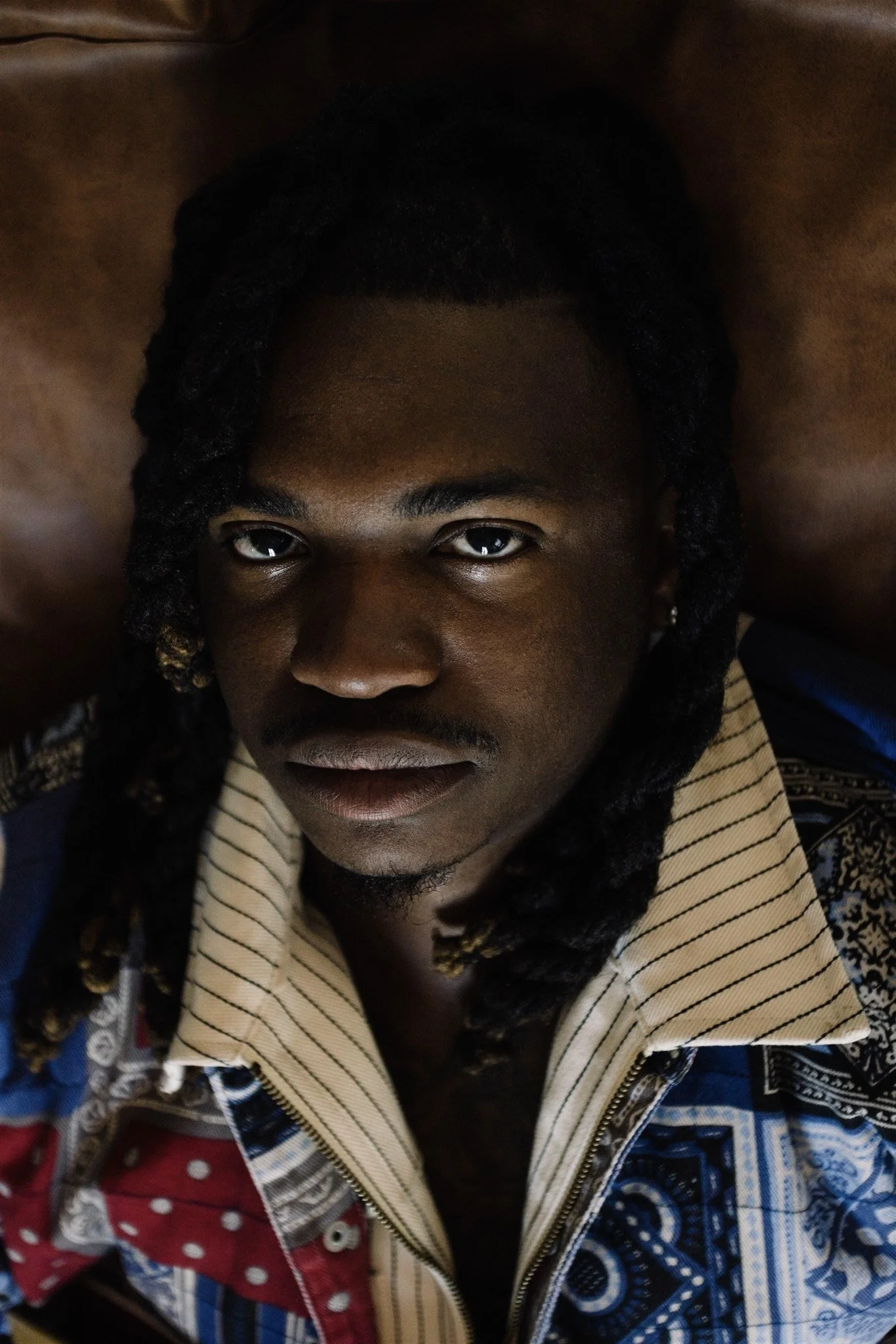 A young man with dark skin and styled dreadlocks looking up at the camera, wearing a colorful patterned jacket with a beige collar.