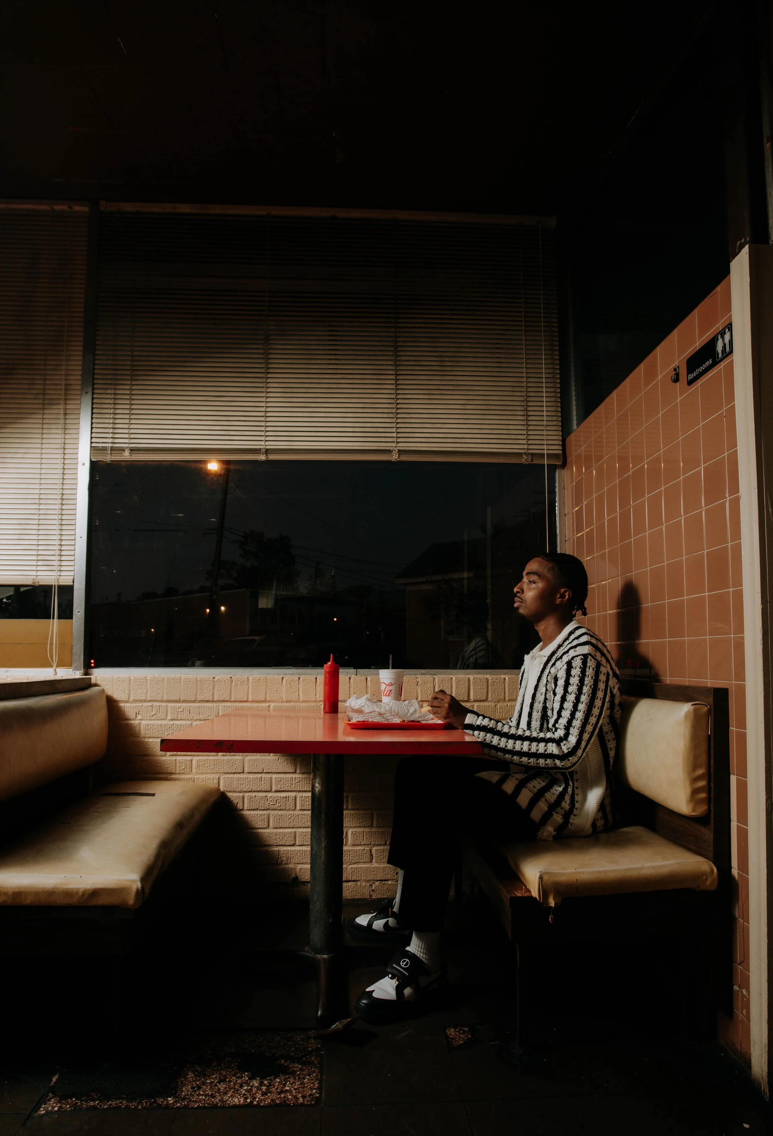 A young man sitting alone at a diner booth during evening, with a red ketchup bottle, a white cup, and food on a red tray in front of him, illuminated by low light.