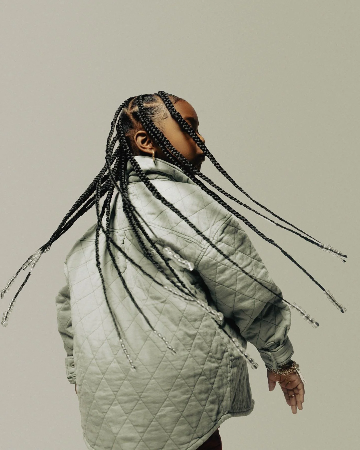 A person with braided hair extending outward, wearing a light gray quilted jacket, facing sideways against a plain background.