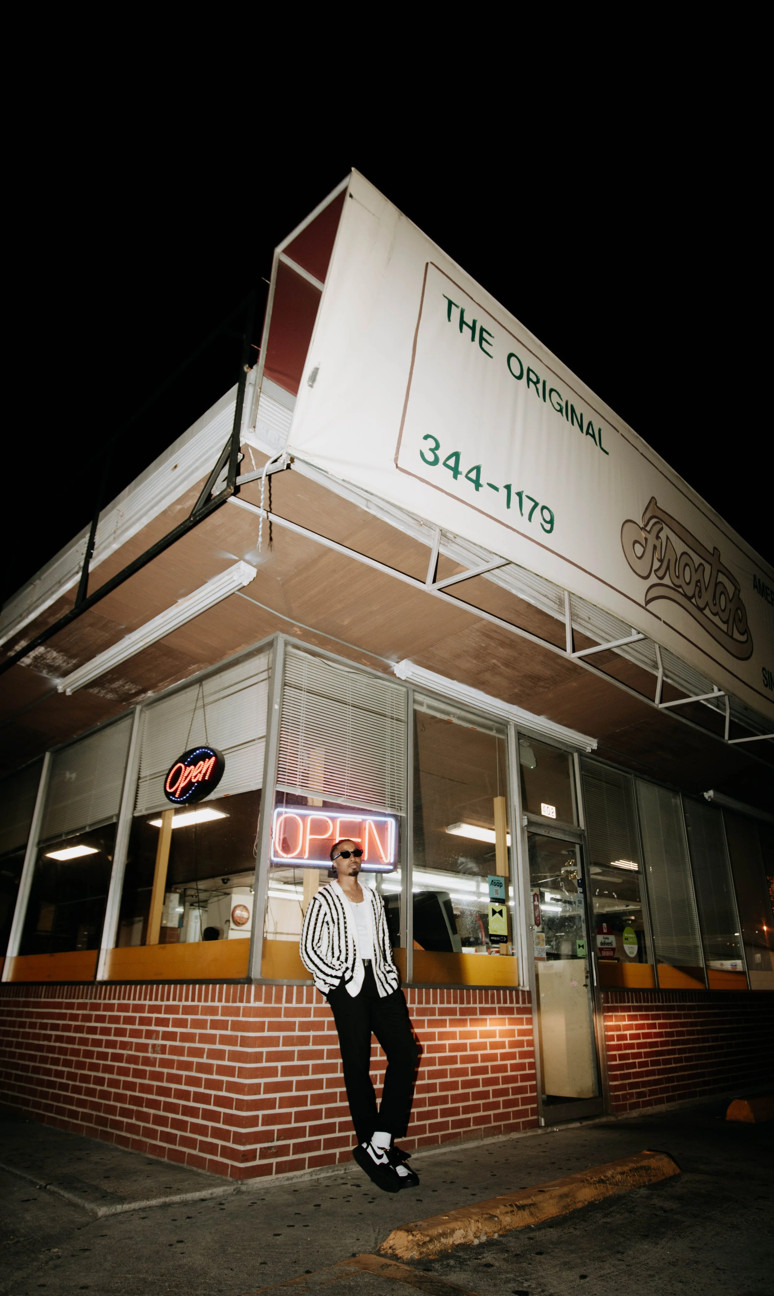 A man in sunglasses and a striped jacket standing outside a fast food restaurant with neon and illuminated "Open" signs at night. The restaurant has a brick base and large glass windows.