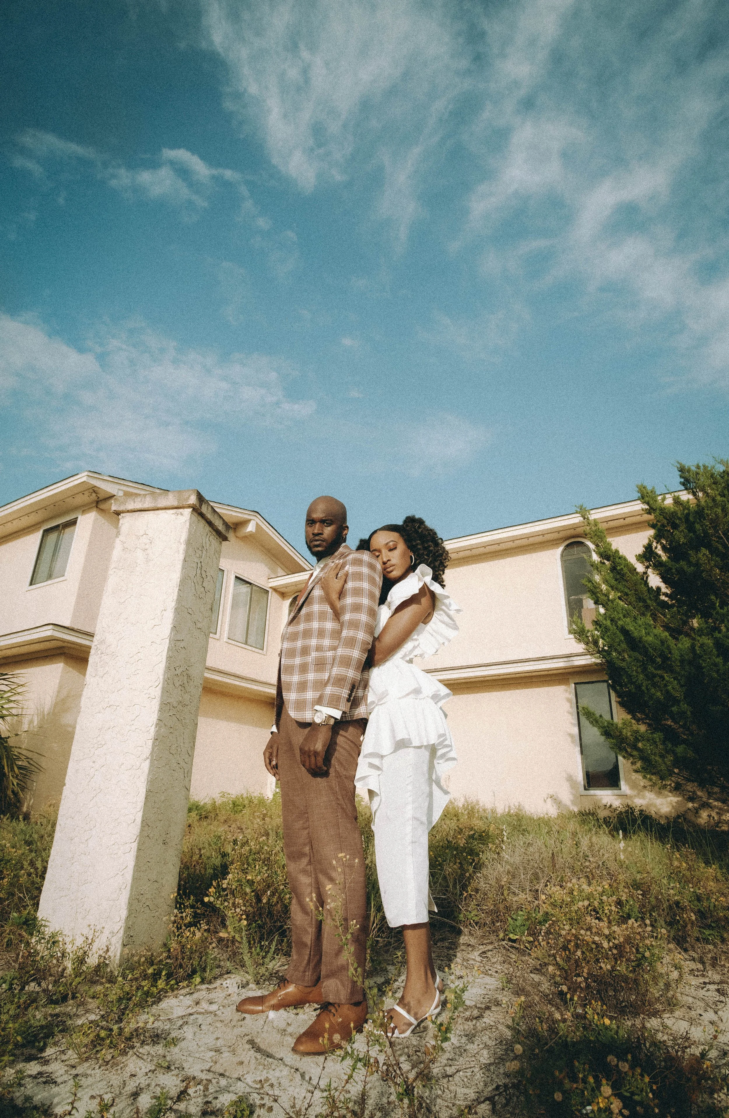 A man in a brown plaid suit and a woman in a white ruffled dress standing outside a house under a cloudy blue sky.