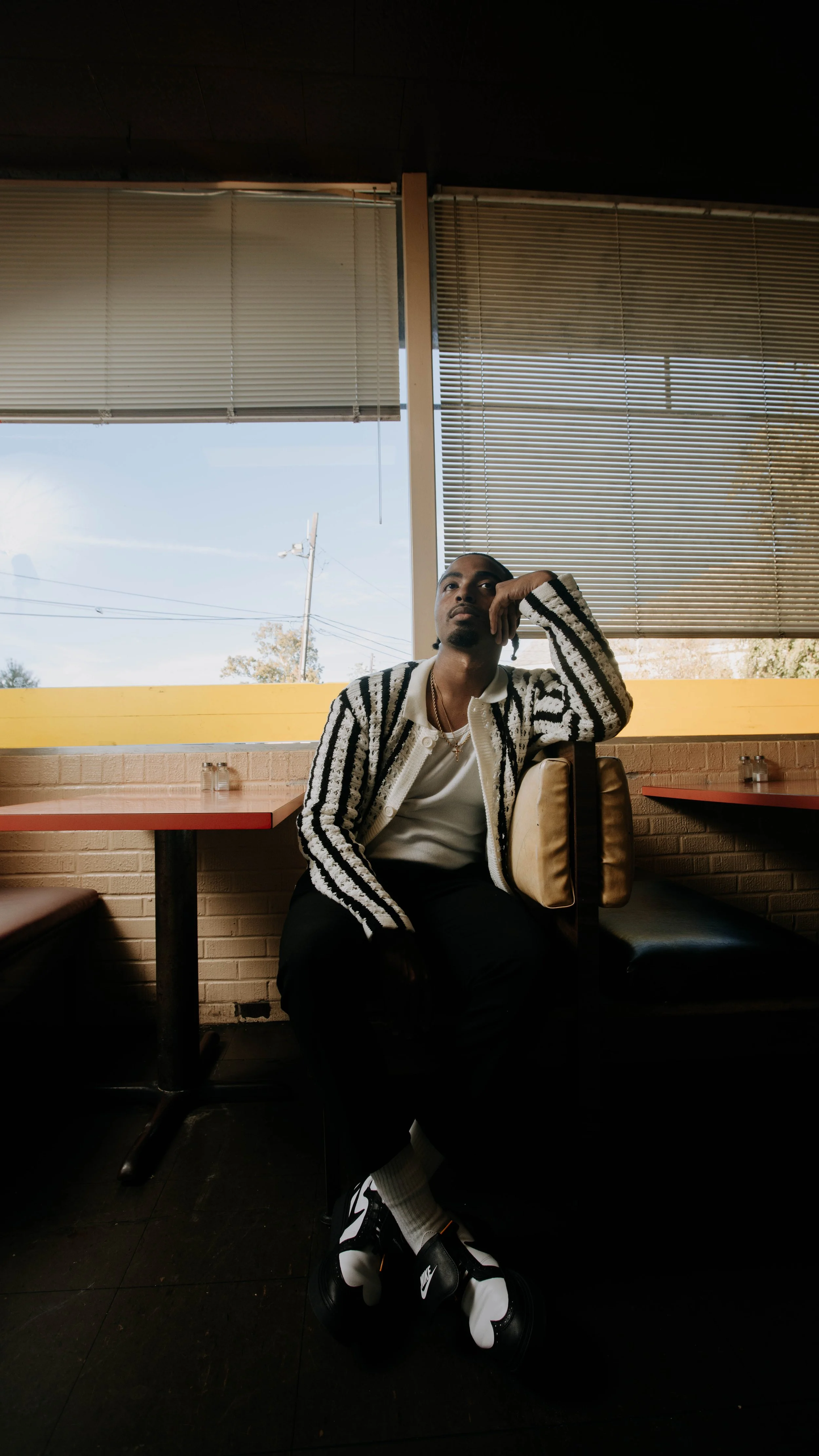 A man sitting in a restaurant booth with his head resting on his hand, wearing a black and white striped cardigan, white shirt, black pants, and sneakers.