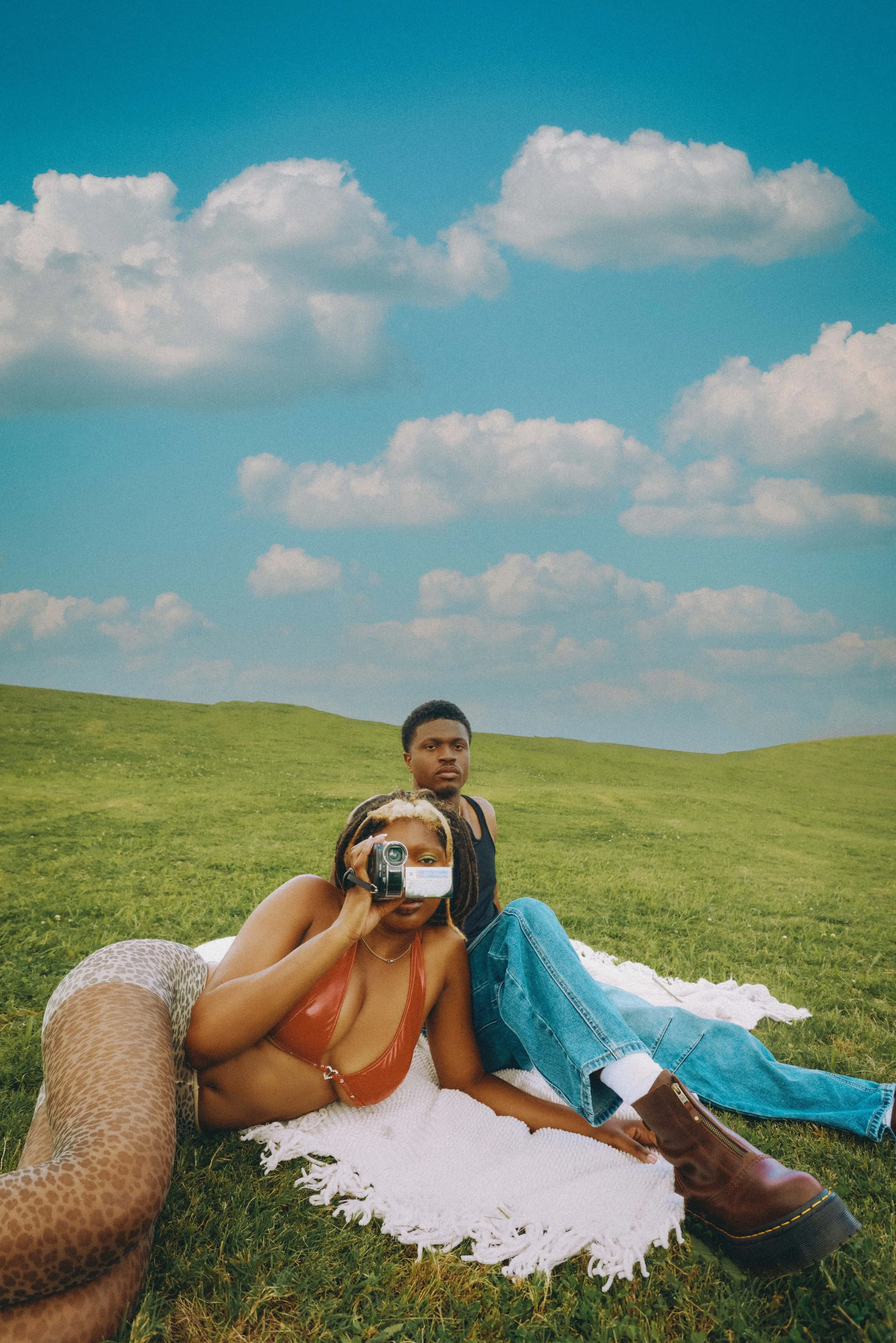 Two individuals lying on a white blanket on the grassy field under a blue sky with clouds, one taking a photo with a small camera and the other looking at the camera. One person is wearing a red top, leopard print pants, and brown boots, while the ot