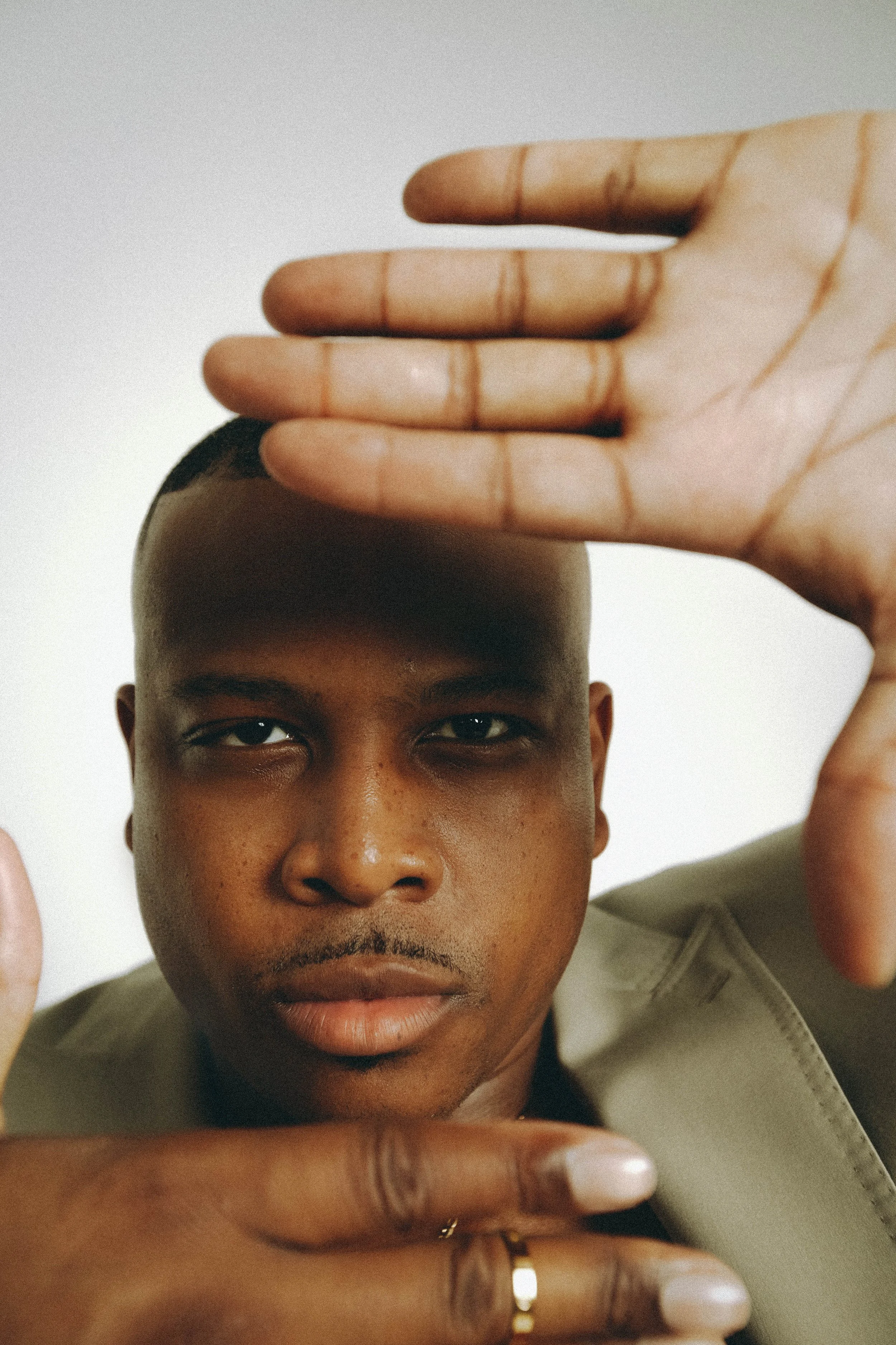 Close-up portrait of a man with short hair, looking directly at the camera, with hands framing his face against a neutral background.