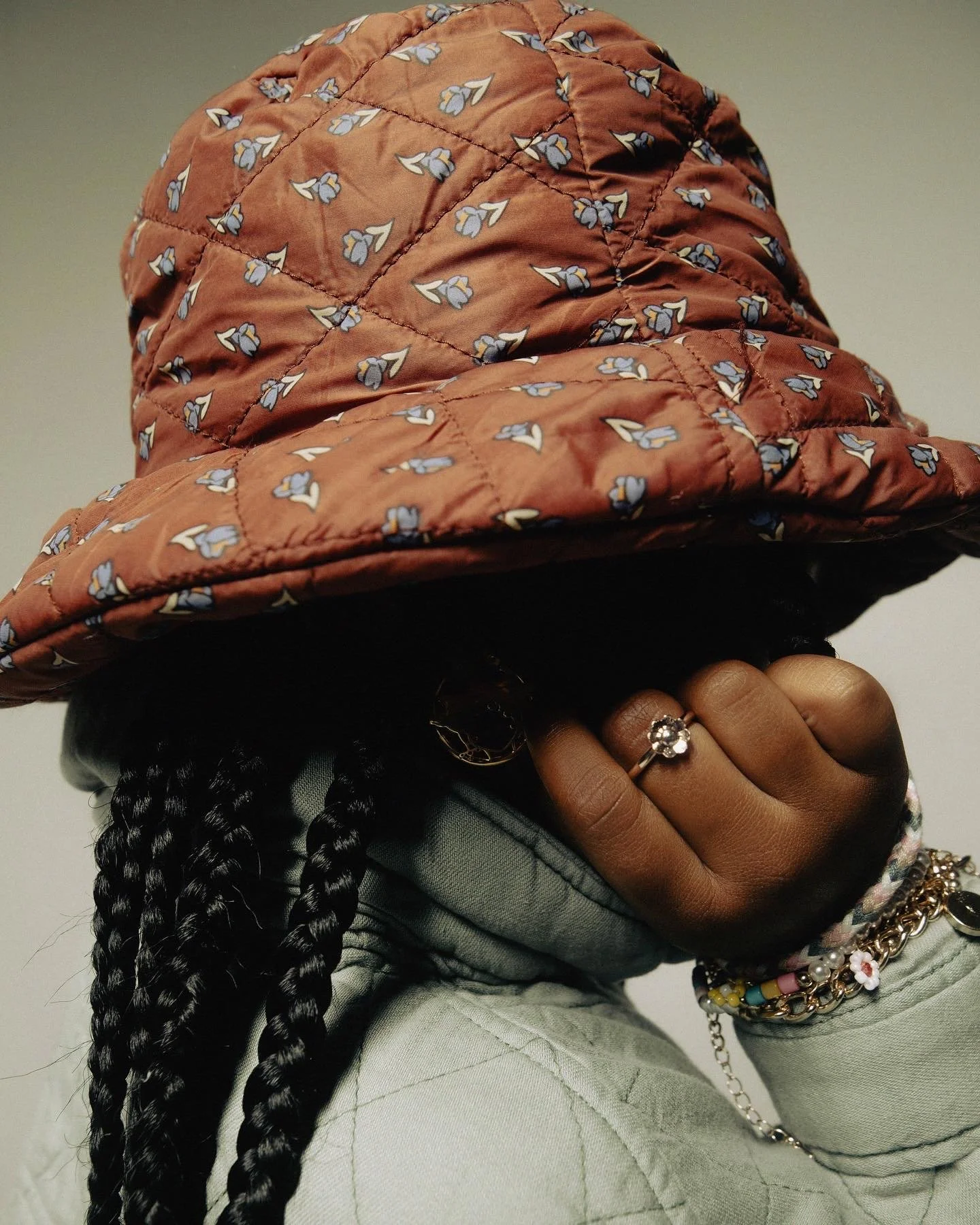 Person wearing a tan quilted hat with blue and white floral pattern, black braids, and jewelry including a flower-shaped ring and bracelets, covering their face with their hand.