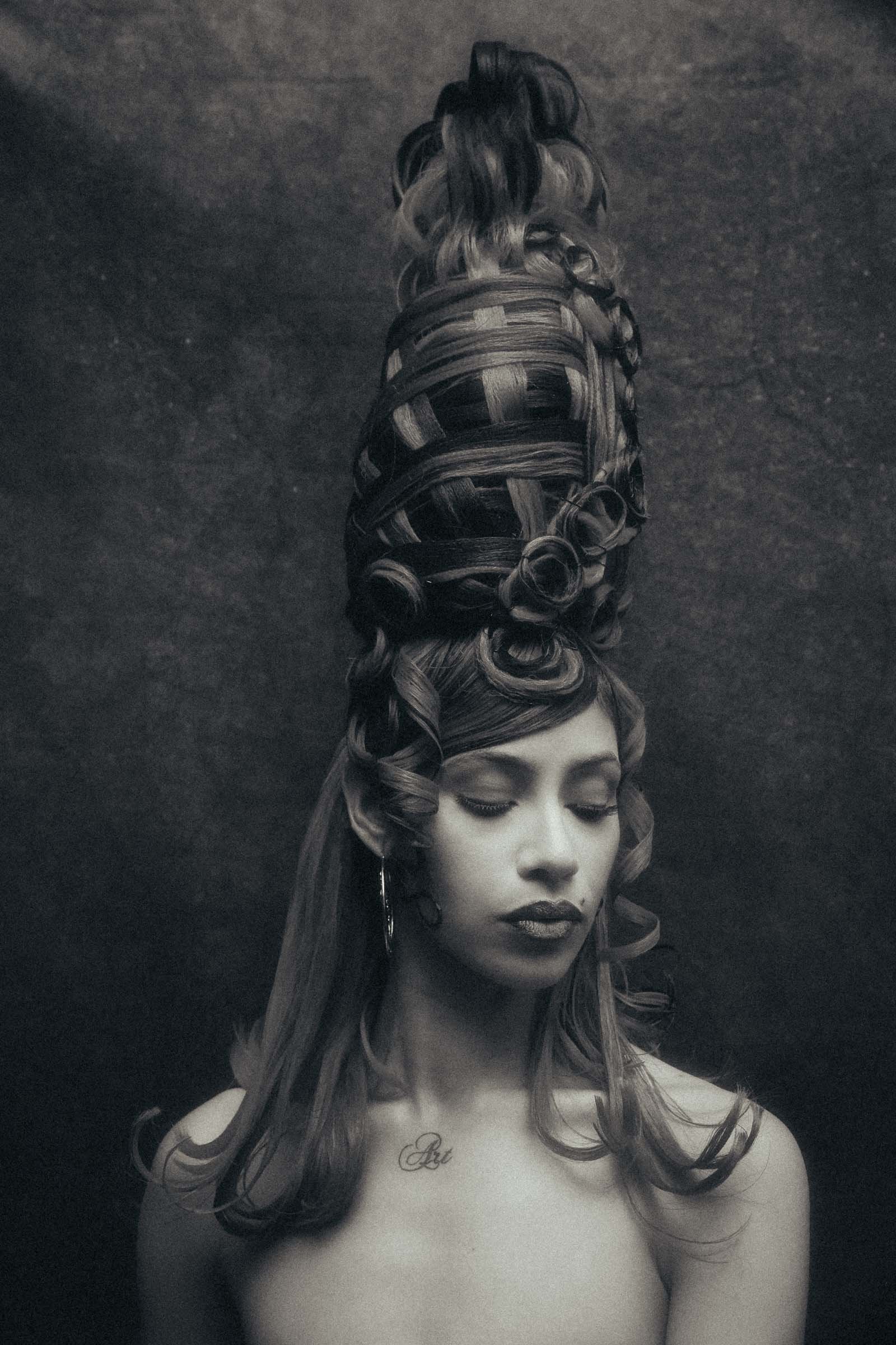 A woman with an elaborate hairstyle featuring curled and woven hair piled high on her head, set against a dark background. She has a calm facial expression with closed eyes and lips, wearing a hoop earring and a tattoo that reads 'Art' on her chest.