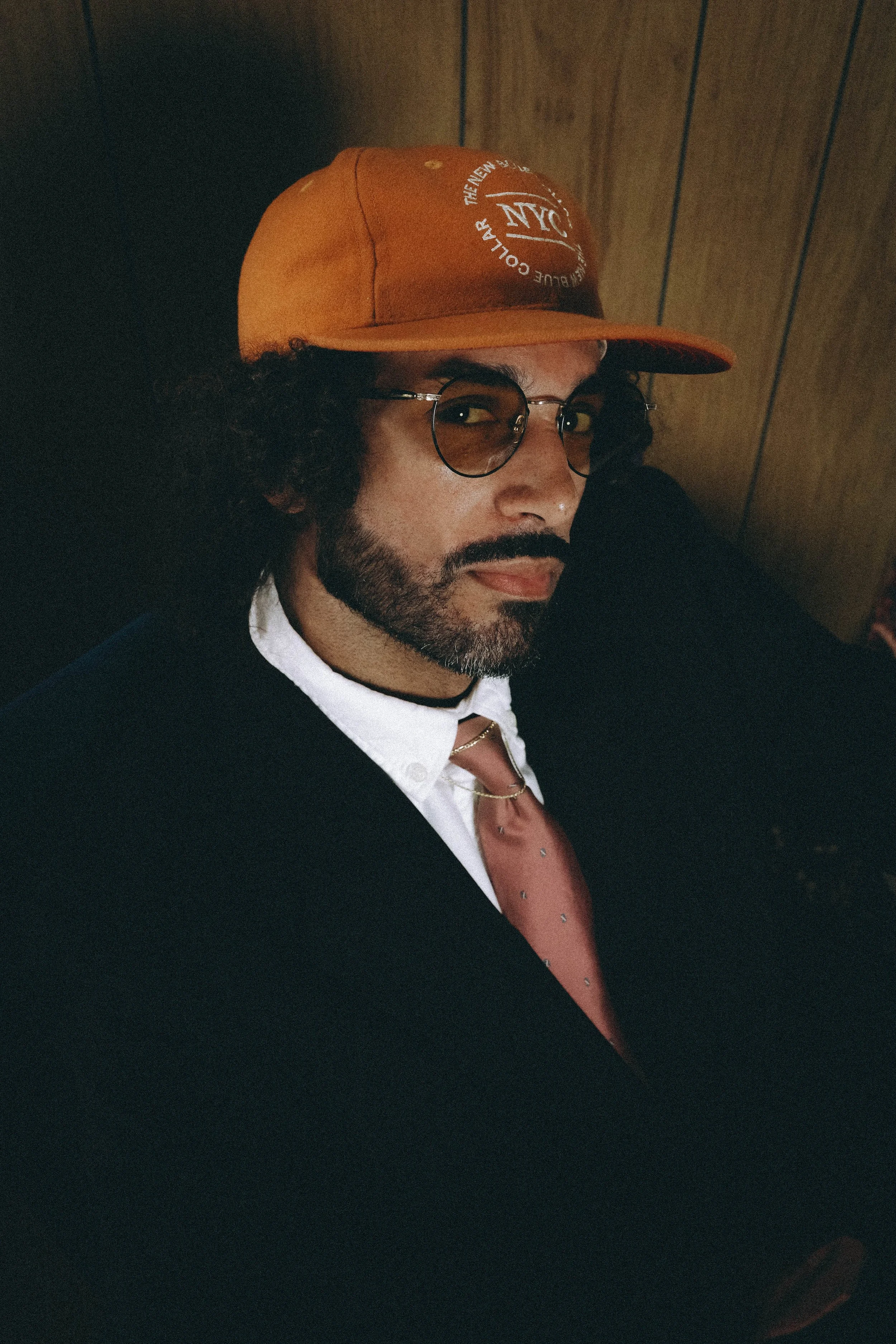 A man with curly hair, glasses, and a beard looking at the camera, wearing an orange cap and a suit with a white shirt and a tie.