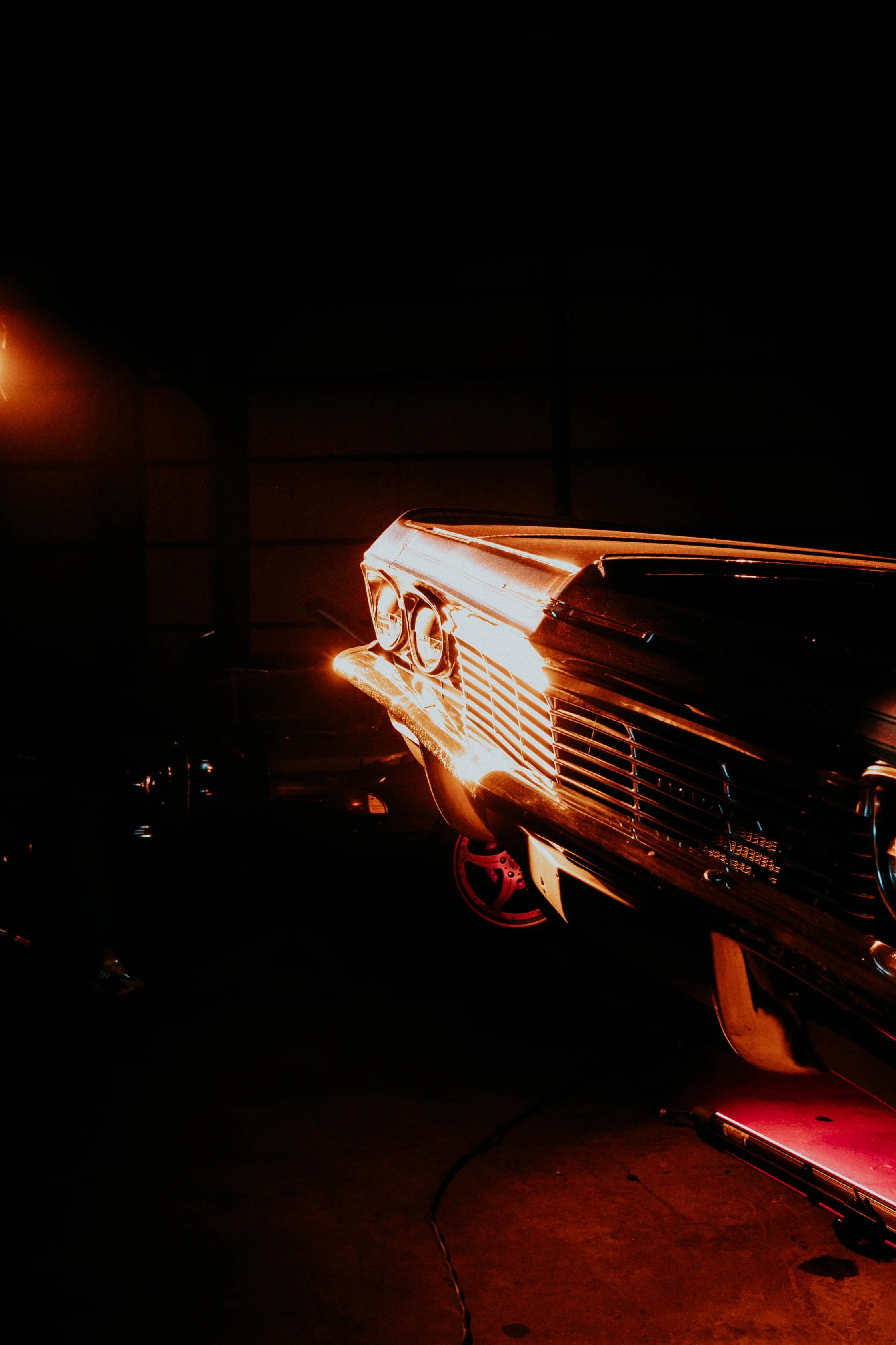 A vintage black car with chrome detailing is illuminated by warm orange lighting in a dark garage or indoor space.
