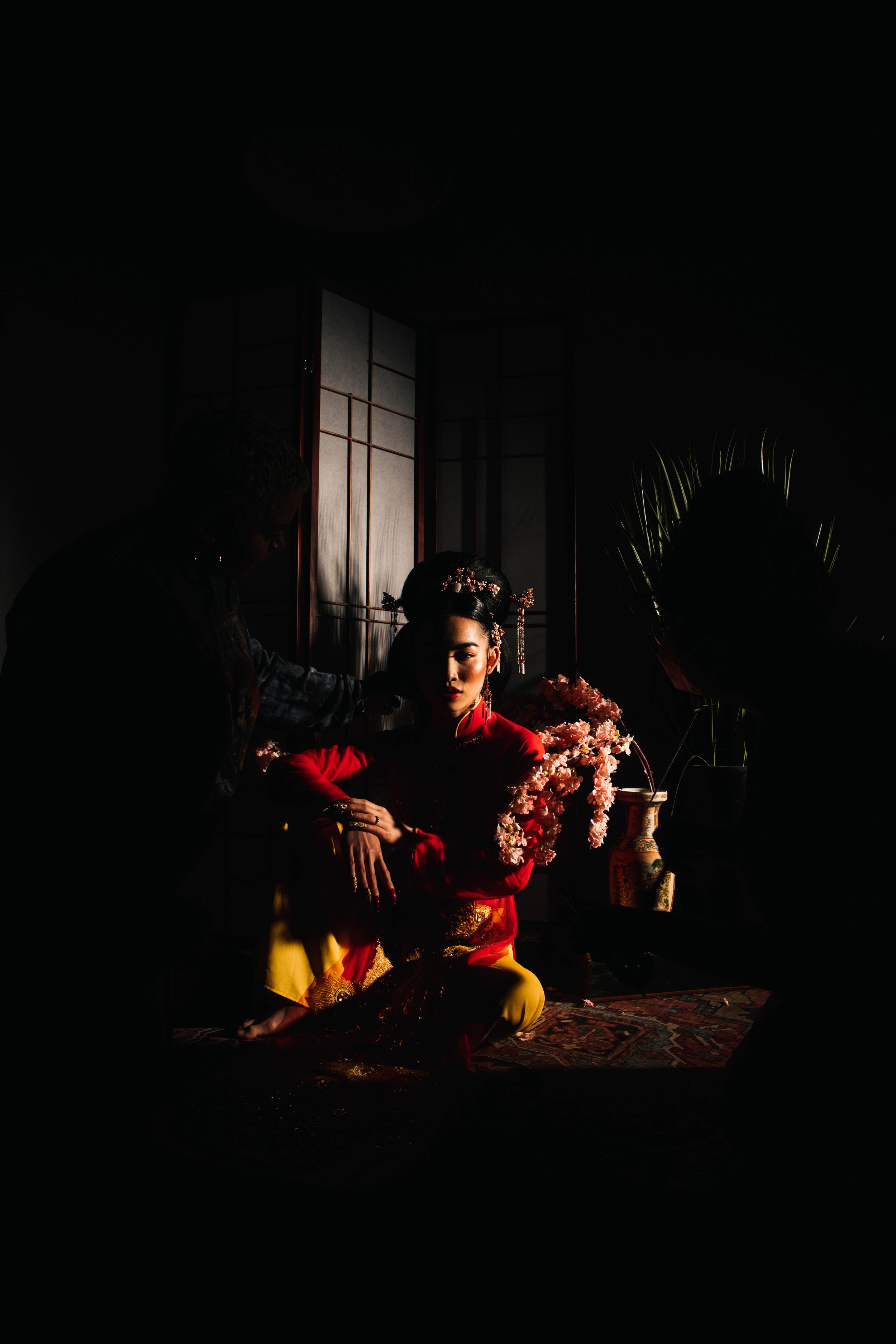 A woman in traditional Asian attire, seated in a dimly lit room with floral decorations and a decorative folding screen.