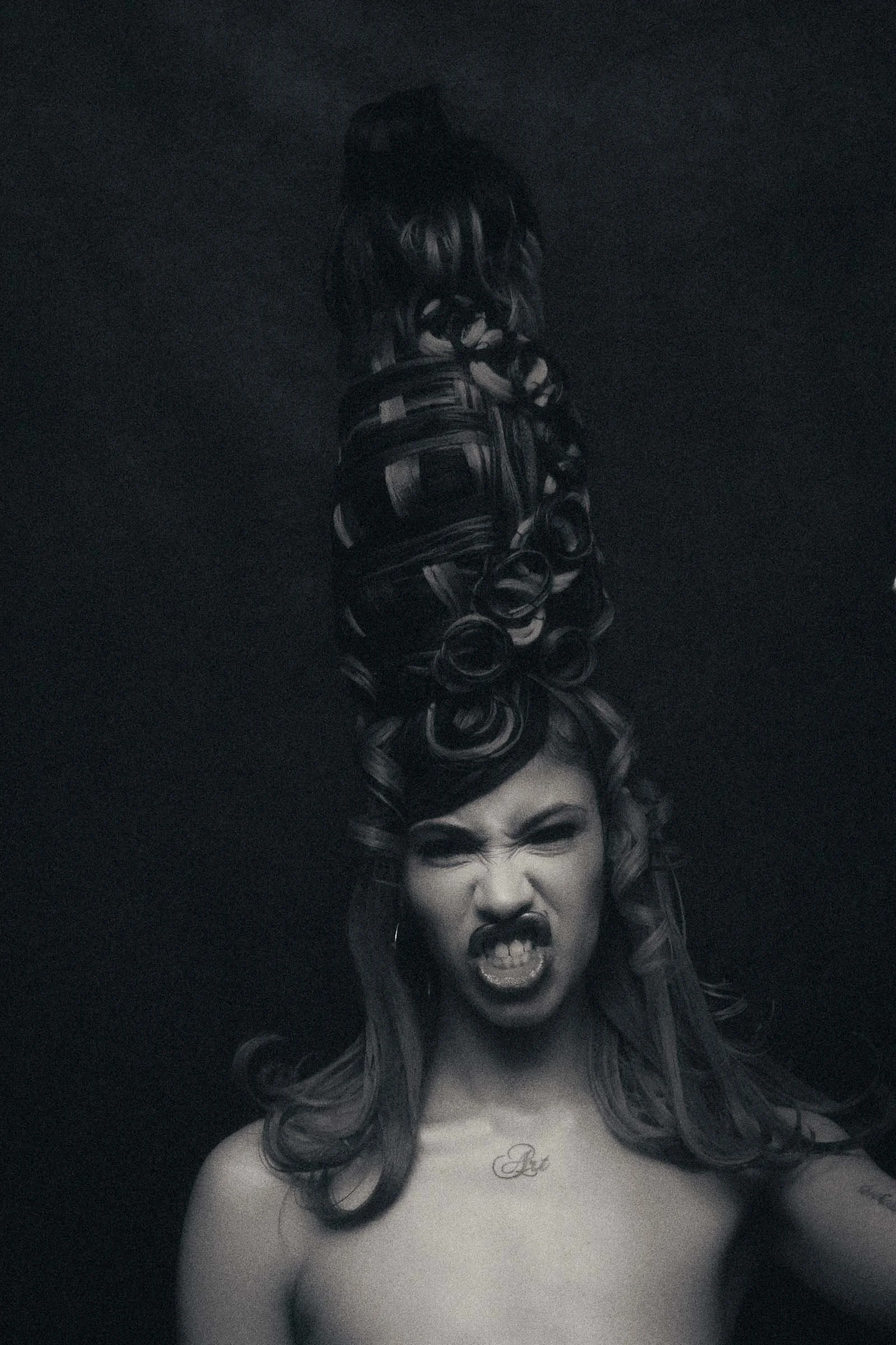 A woman underwater with hair styled into a tall, intricate updo. She is making a fierce facial expression with her teeth bared and one eye squinting.
