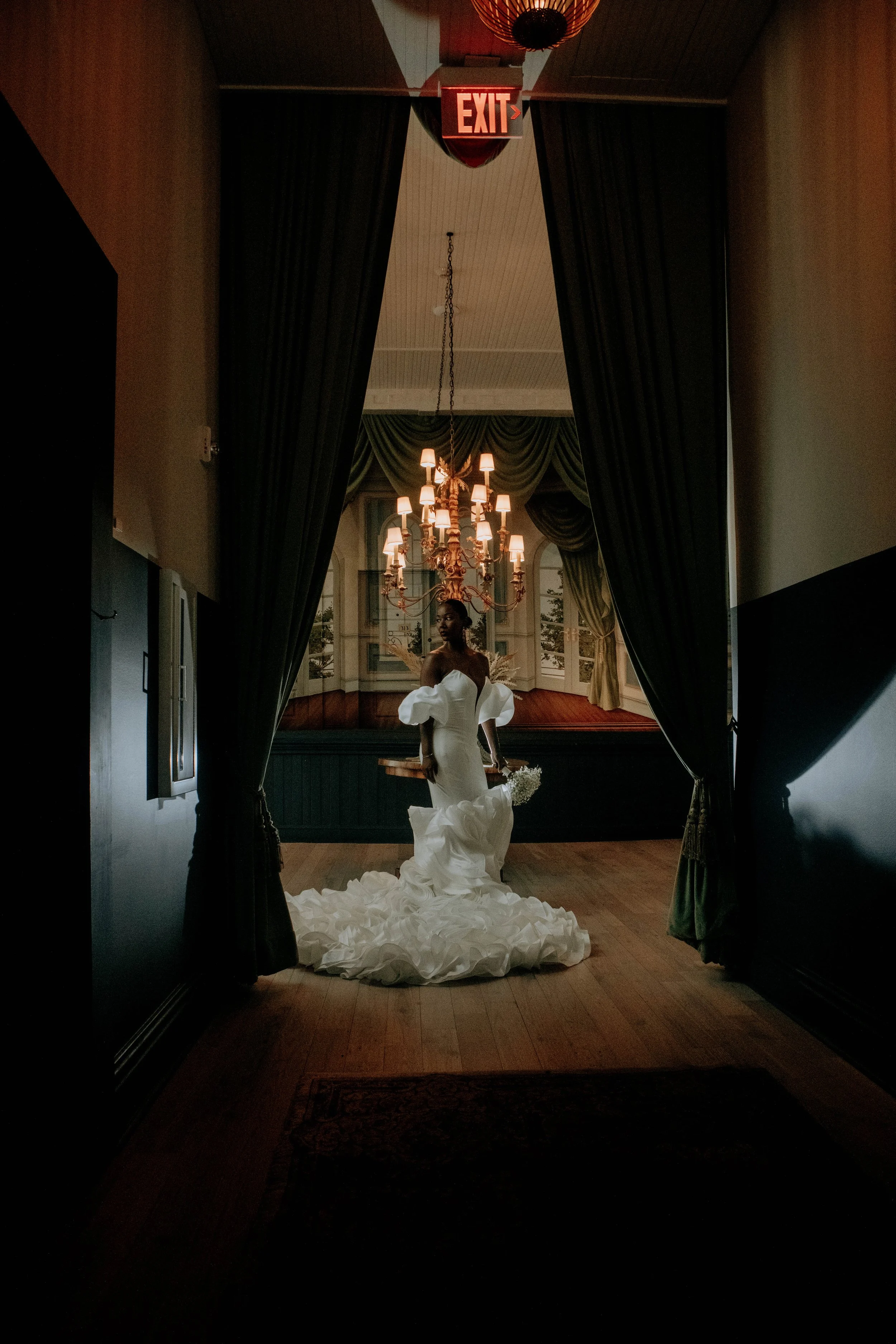 A woman in a white wedding gown standing in a dimly lit room framed by dark curtains, with a chandelier and large windows in the background.