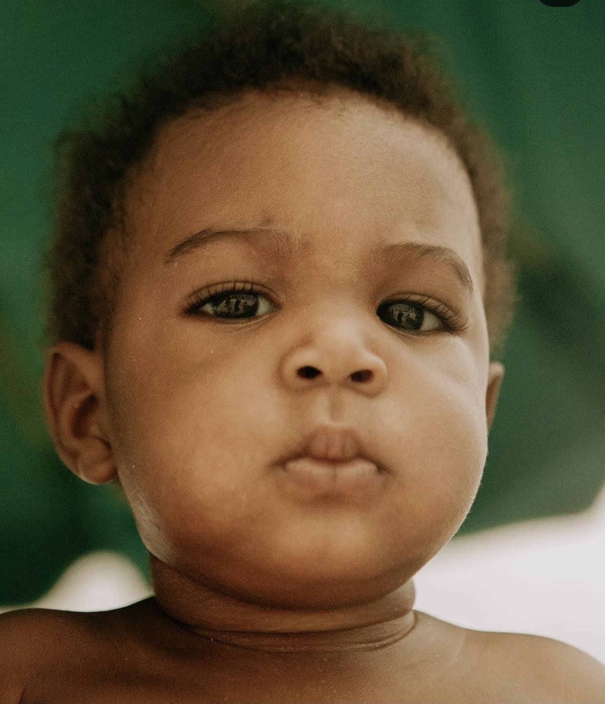 A close-up photo of a young child with tan skin and curly hair, looking directly at the camera with a neutral expression.