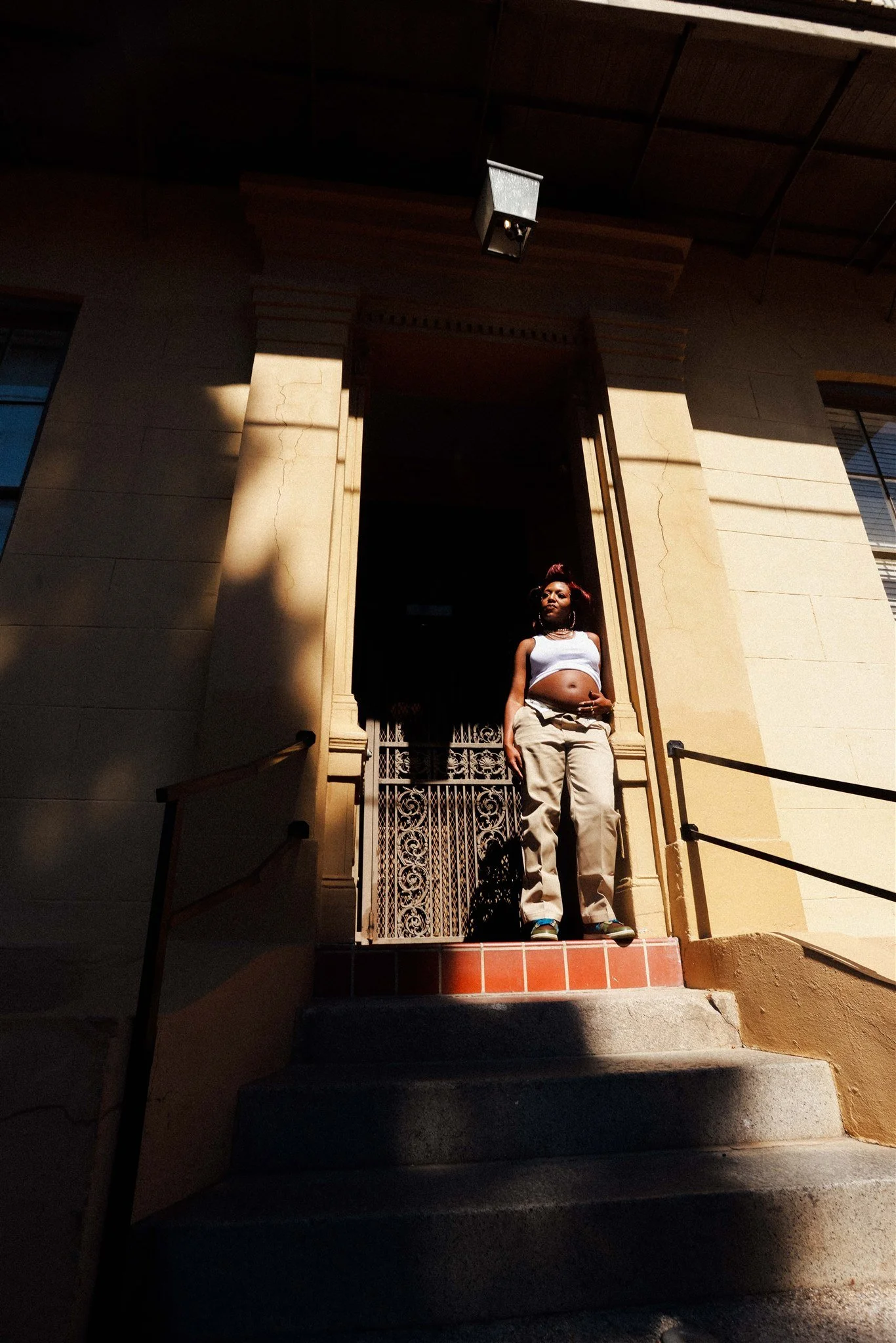 A woman standing on steps outside a building with a decorative metal gate at the entrance, wearing a white crop top and beige pants, with sunlight casting shadows on the wall.