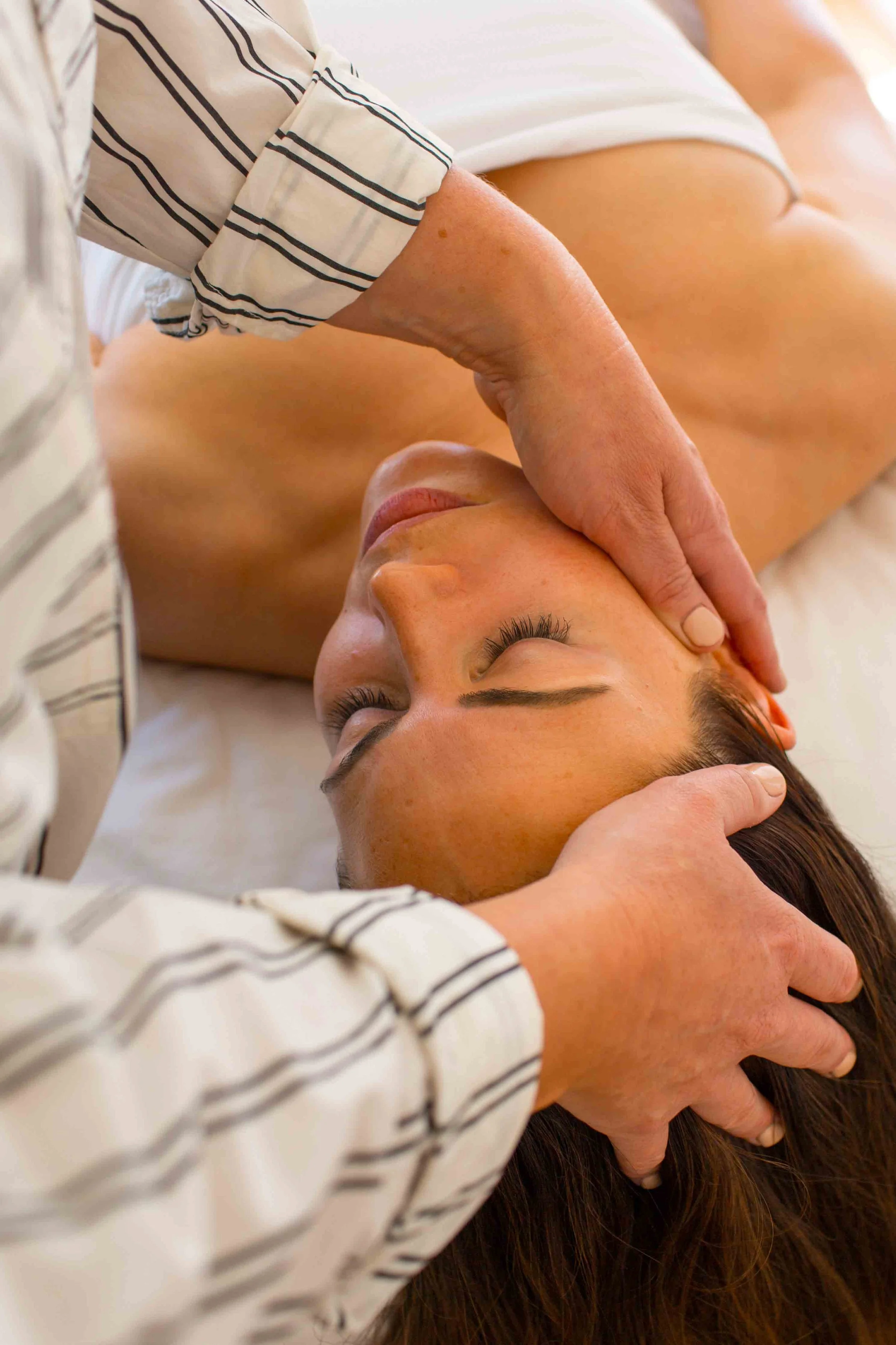 A person receiving a facial massage while lying down, with someone else gently pressing on their forehead and scalp, wearing a white and black striped shirt.