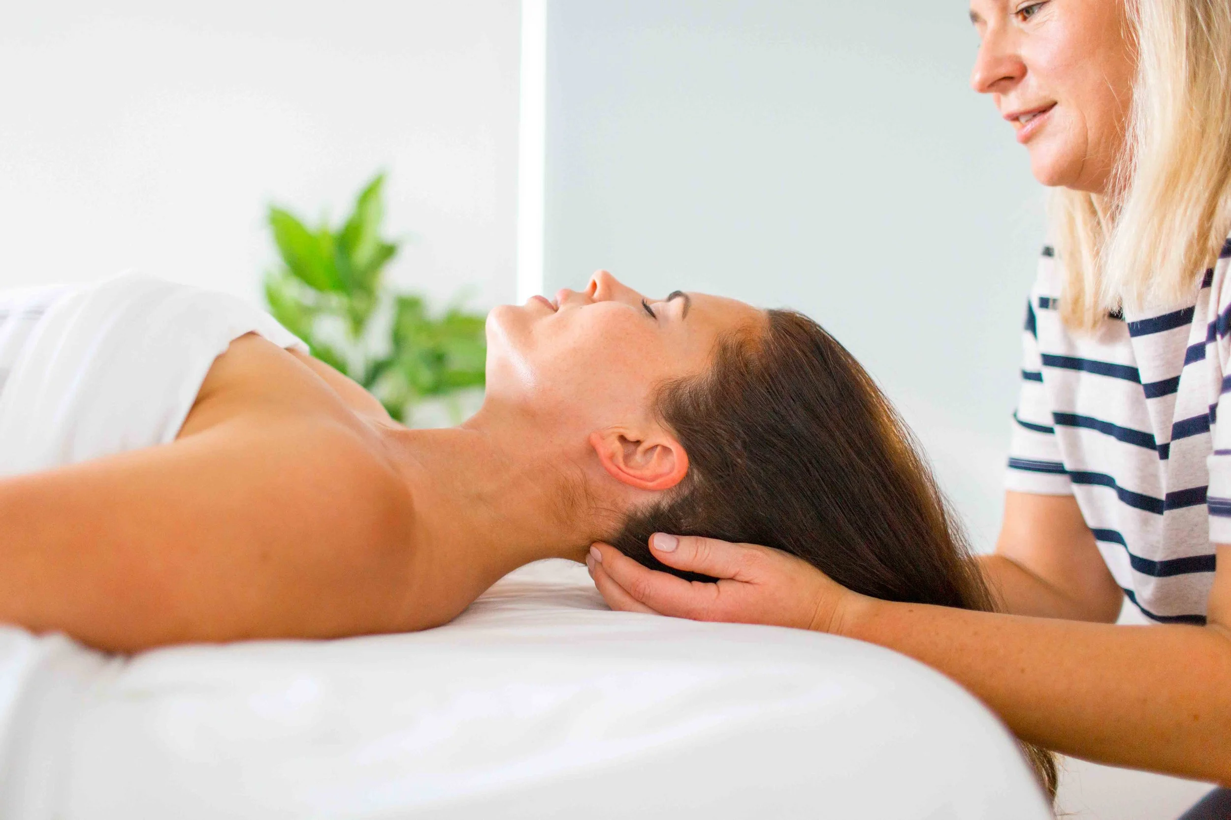 A woman receiving a massage from a massage therapist in a calm, bright room with a green plant in the background.