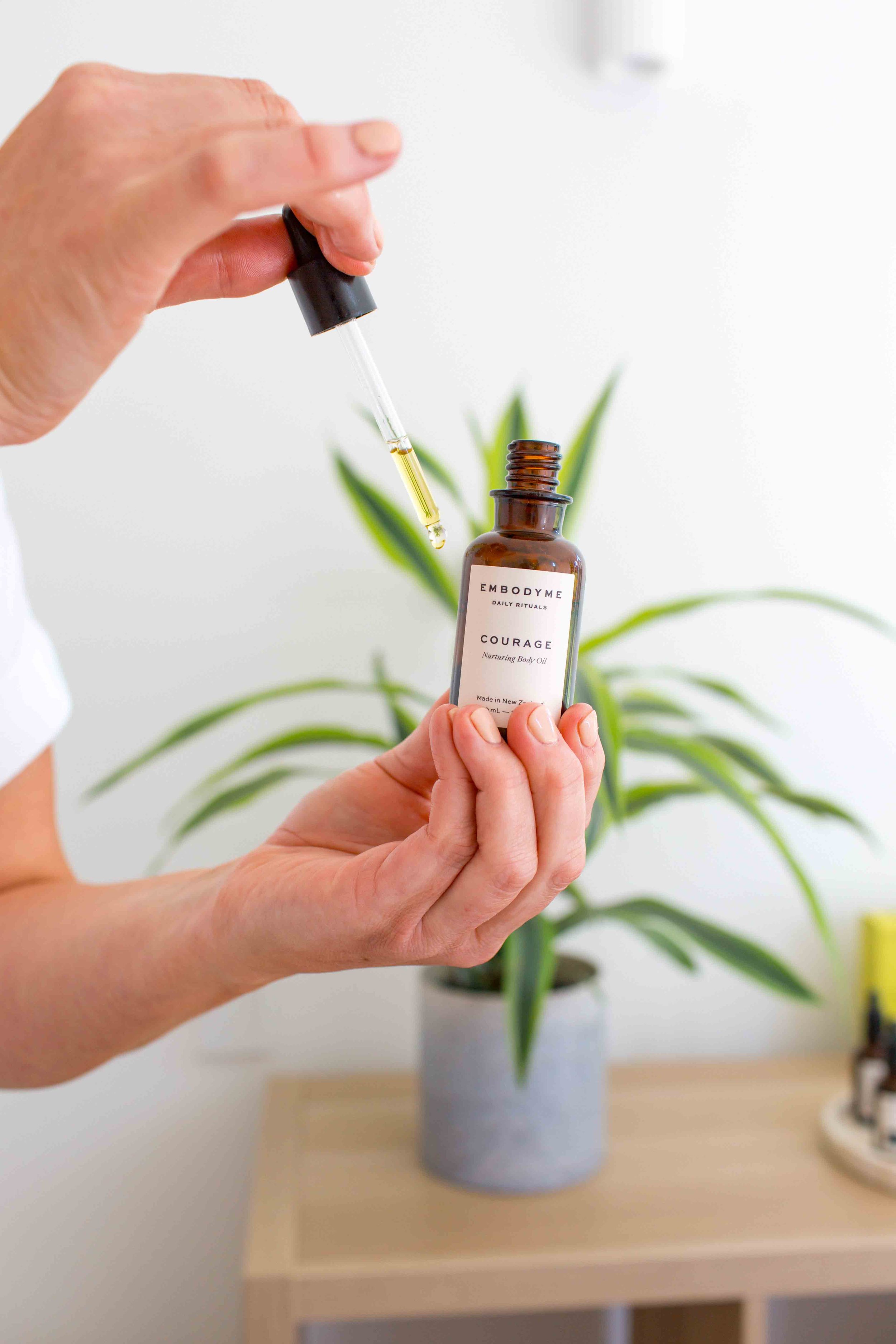 Person holding a small brown bottle labeled 'Embodyme Courage Nurturing Body Oil' and using a dropper to dispense oil.