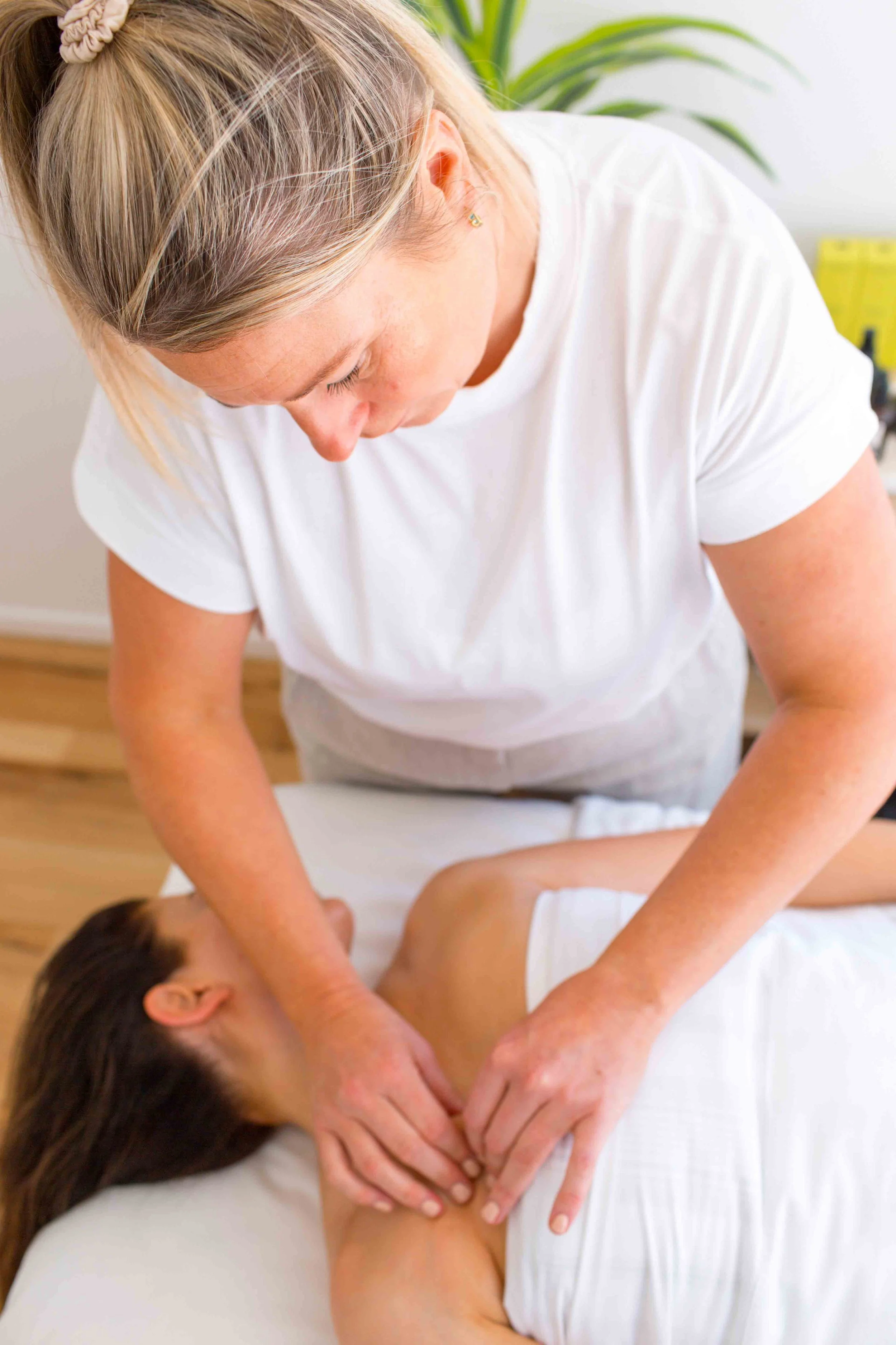 A woman receives a massage while lying on a massage table. The masseuse, wearing a white shirt, is applying pressure to the client's back with her hands. The client has brown hair and is covered with a white towel. There is a green plant in the background.