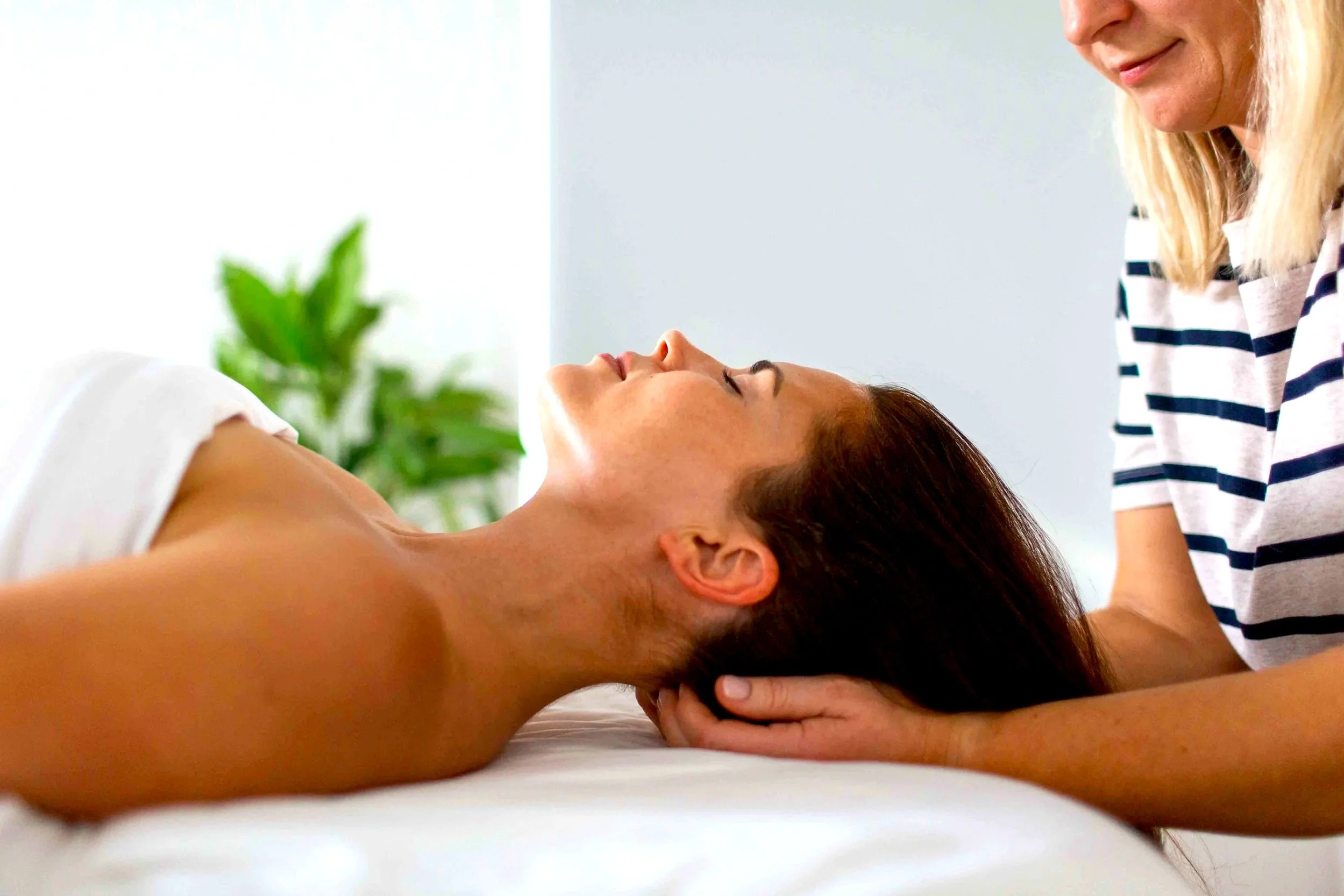 A woman receiving a massage while lying on her back with her eyes closed, and a massage therapist gently supporting her head with one hand.